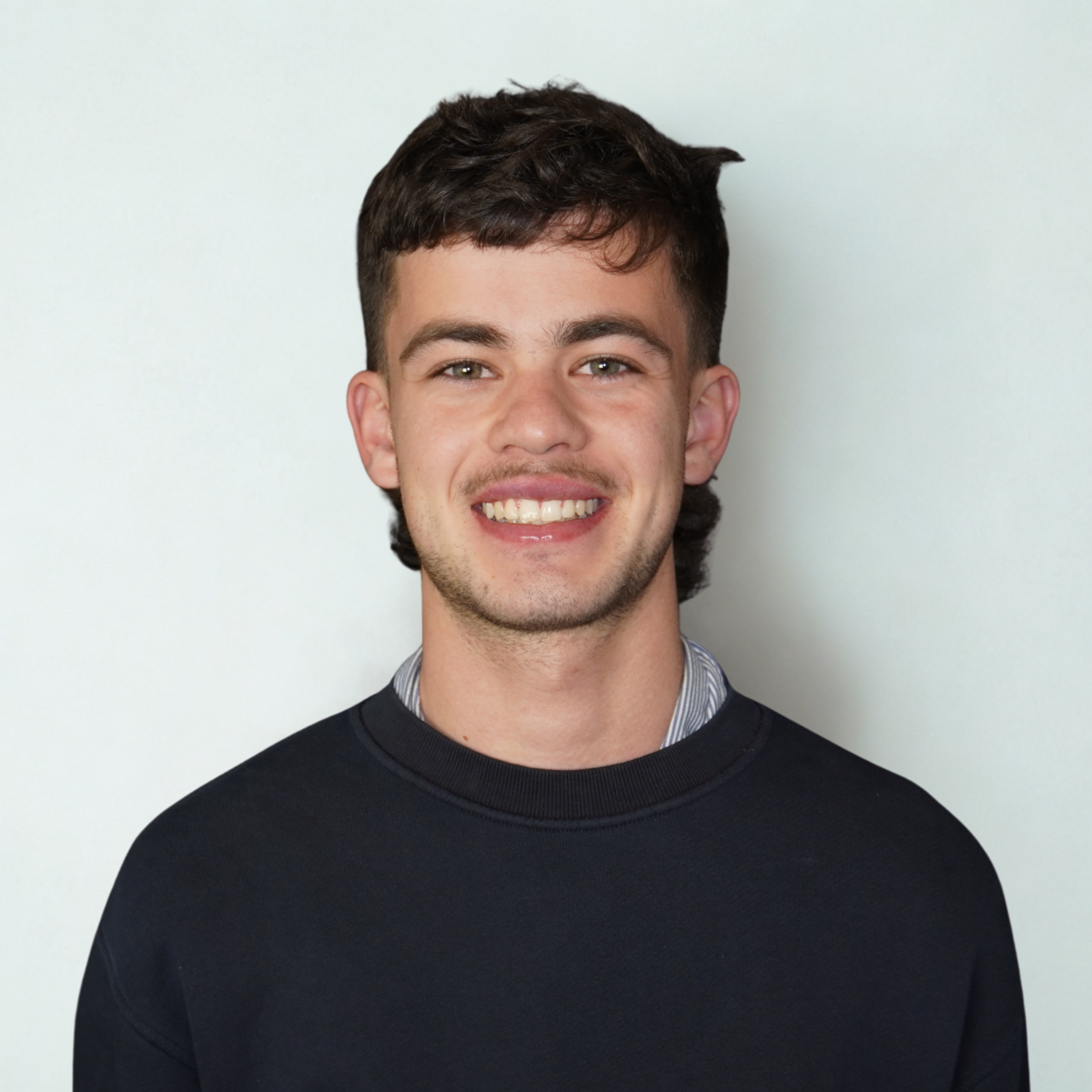 Smiling young man with brown hair wearing a black sweater and a collared shirt underneath, standing against a plain light background.