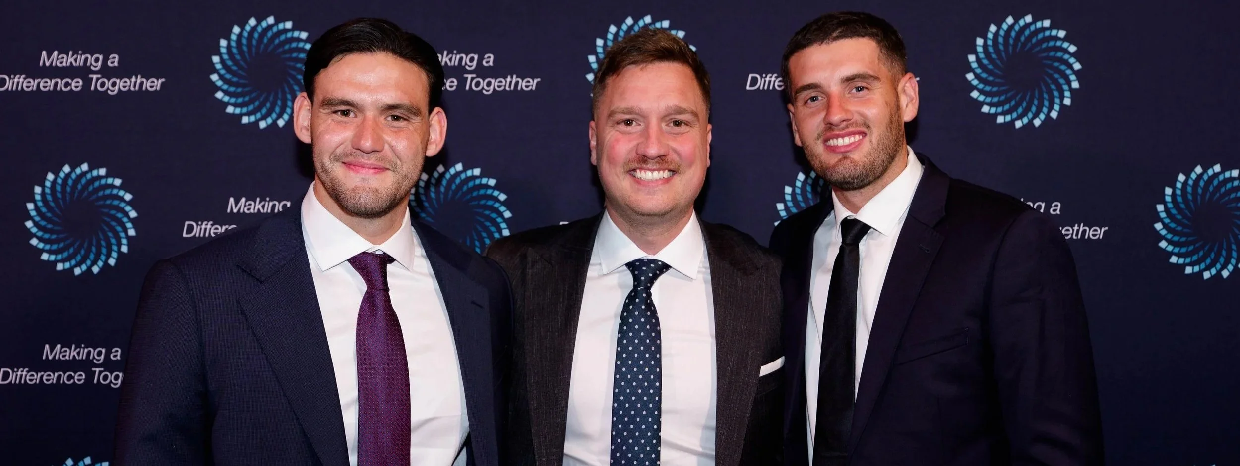 Three men in formal suits smiling for a photo at an event with a dark backdrop featuring a logo and the text 'Making a Difference Together'.