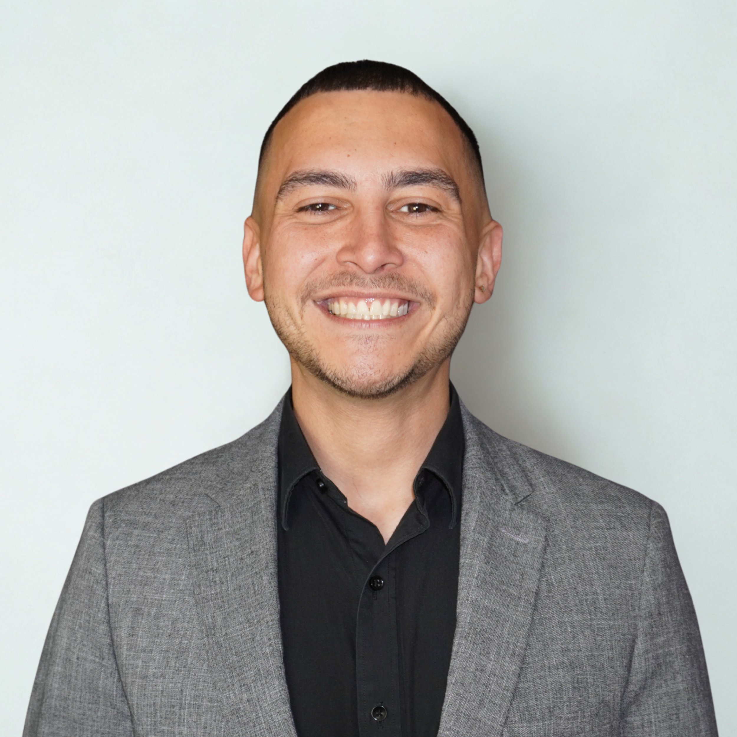 Headshot of a smiling man with short dark hair, wearing a black shirt and gray blazer, standing against a light background.