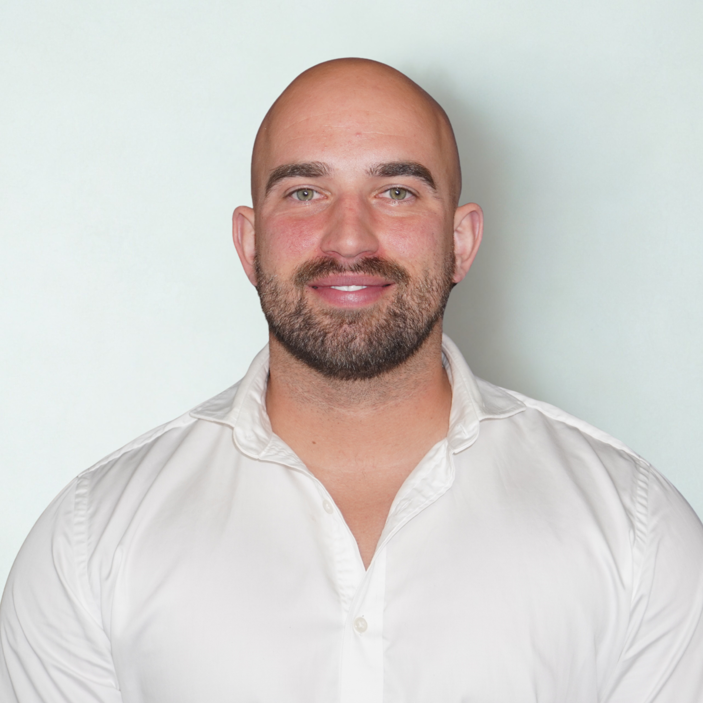 Portrait of a man with a shaved head, full beard, wearing a white button-up shirt, smiling, against a plain light-colored background.