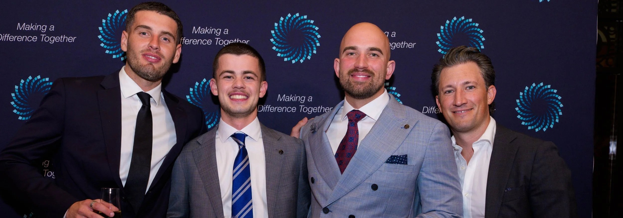 Four men in suits smiling at an event with a dark backdrop that has a logo and the phrase "Making a Difference Together."