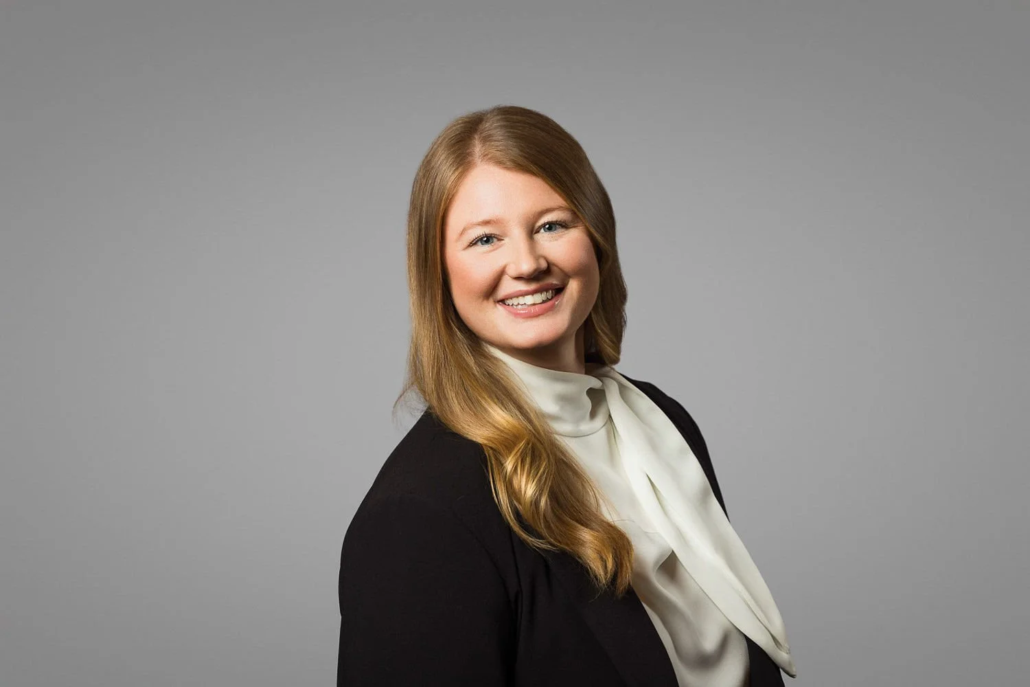 A professional woman with long blonde hair, wearing a black blazer and a white blouse with a bow, standing against a plain gray background.