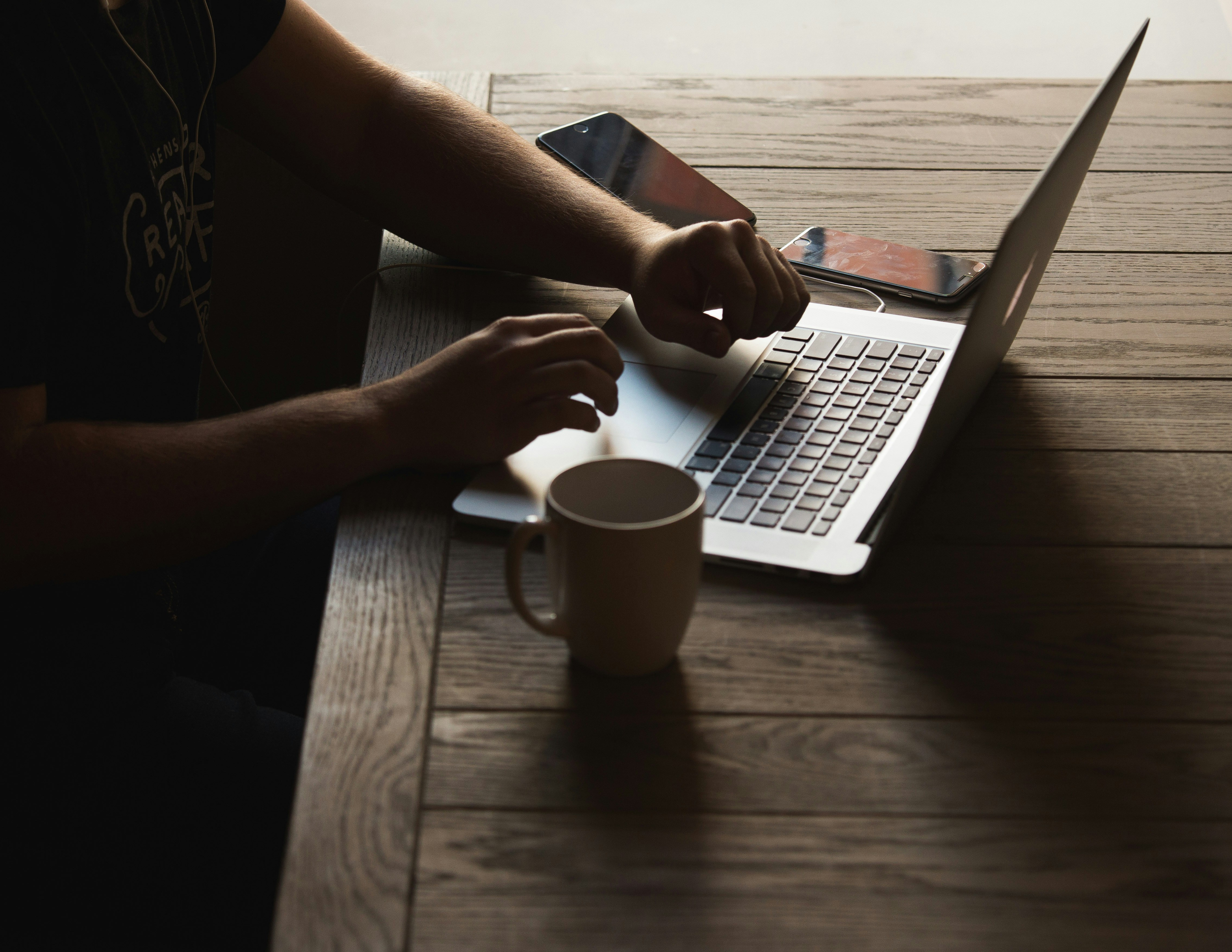 Person working on a laptop at a wooden table with a coffee mug and two smartphones nearby.