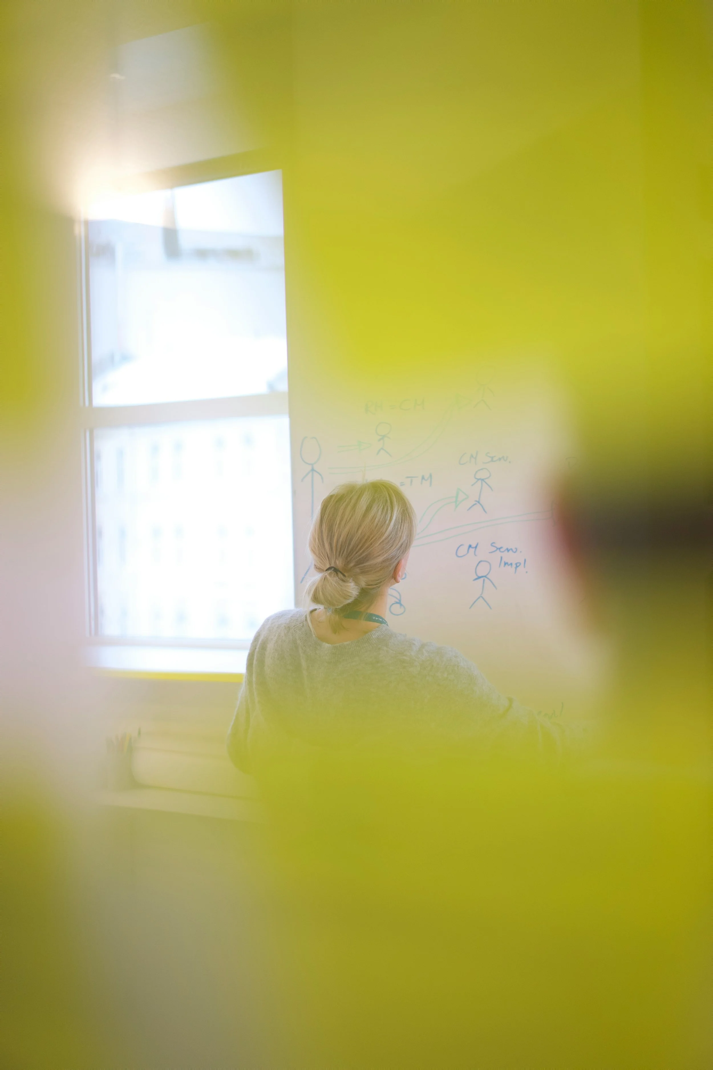 Person with blonde hair writing on a whiteboard, seen through a yellow-tinted window or barrier.