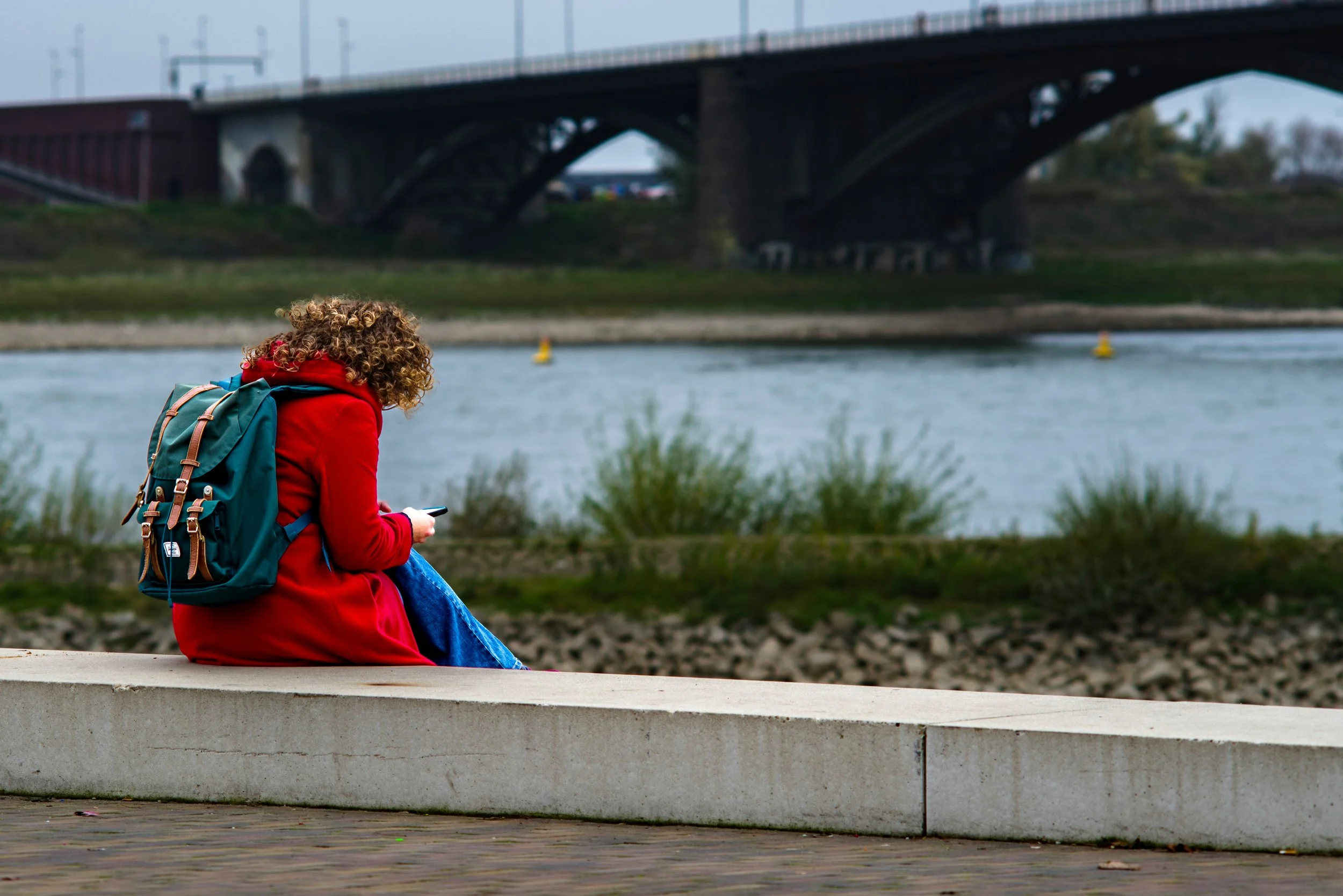 A person with curly hair, wearing a red coat and blue jeans, sitting on a concrete bench by a river, looking at a mobile phone. They have a backpack on their back. In the background, there is a bridge over the river.