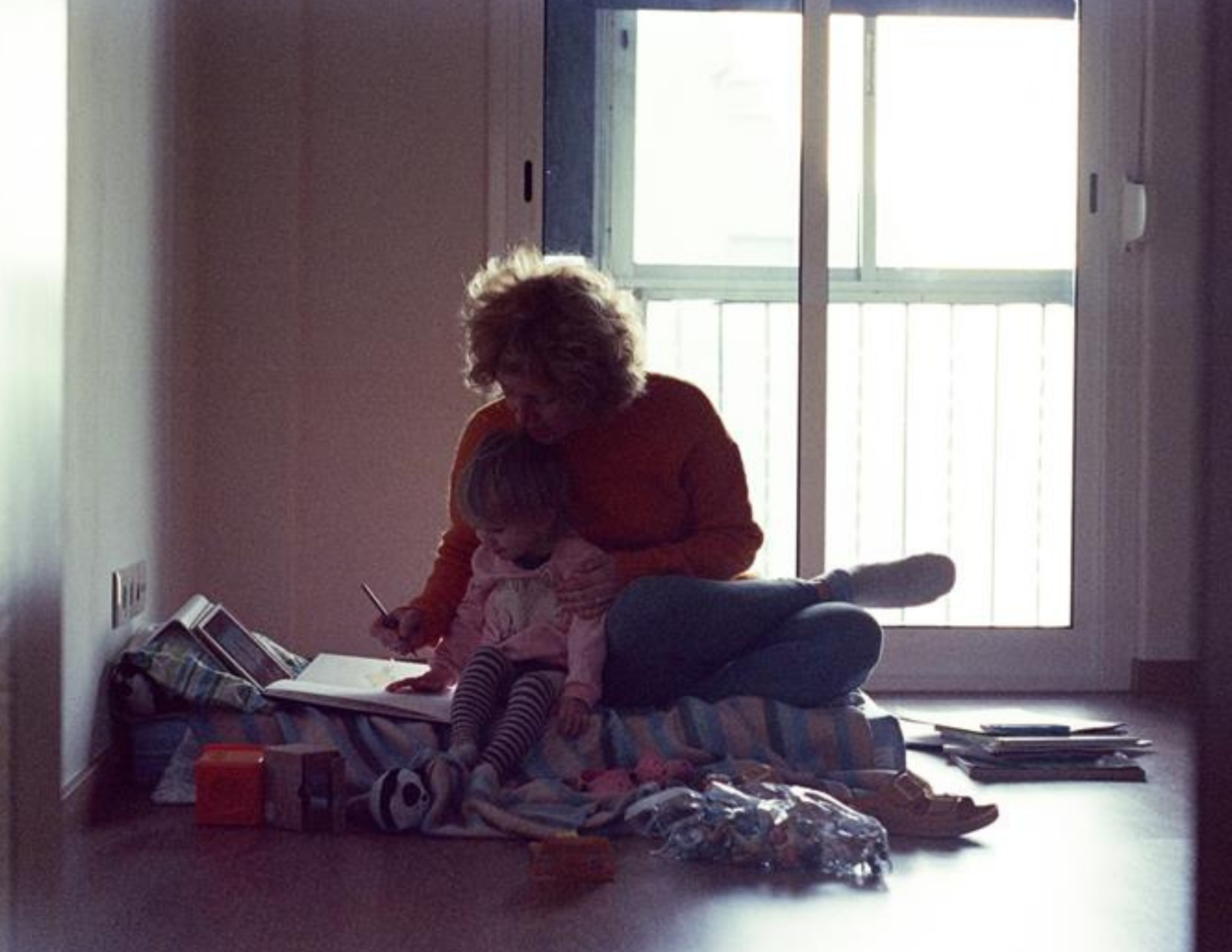 A woman sits on the floor with a young girl, reading a book together near a sliding glass door. The woman is wearing a red top, and the girl is dressed in striped leggings.
