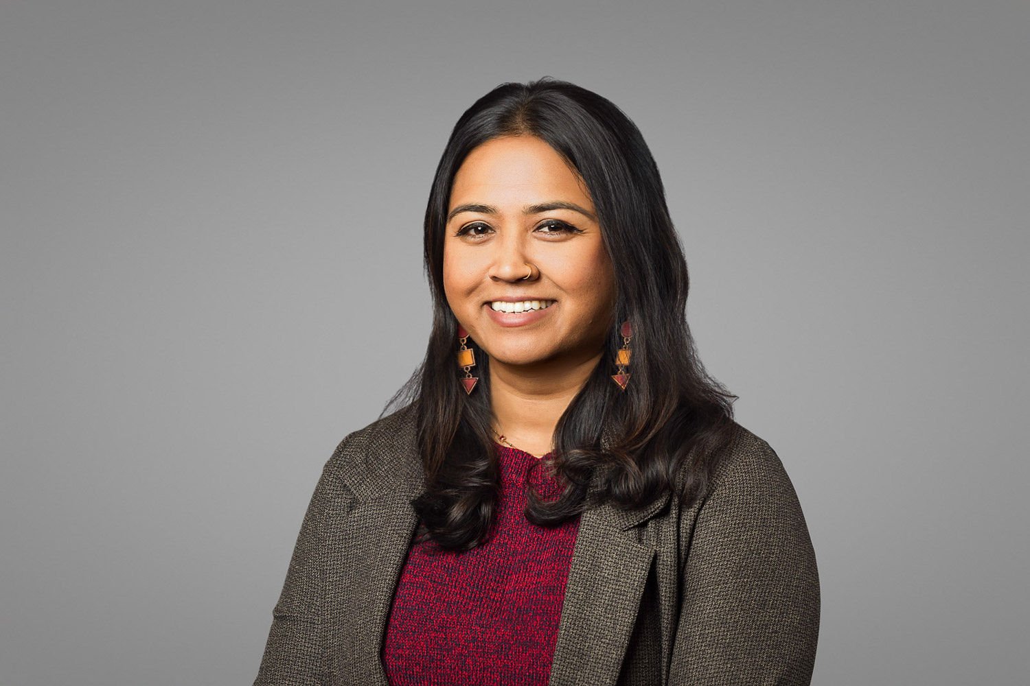 A professional woman with long dark hair, wearing a blazer and red top, smiling at the camera against a plain gray background.