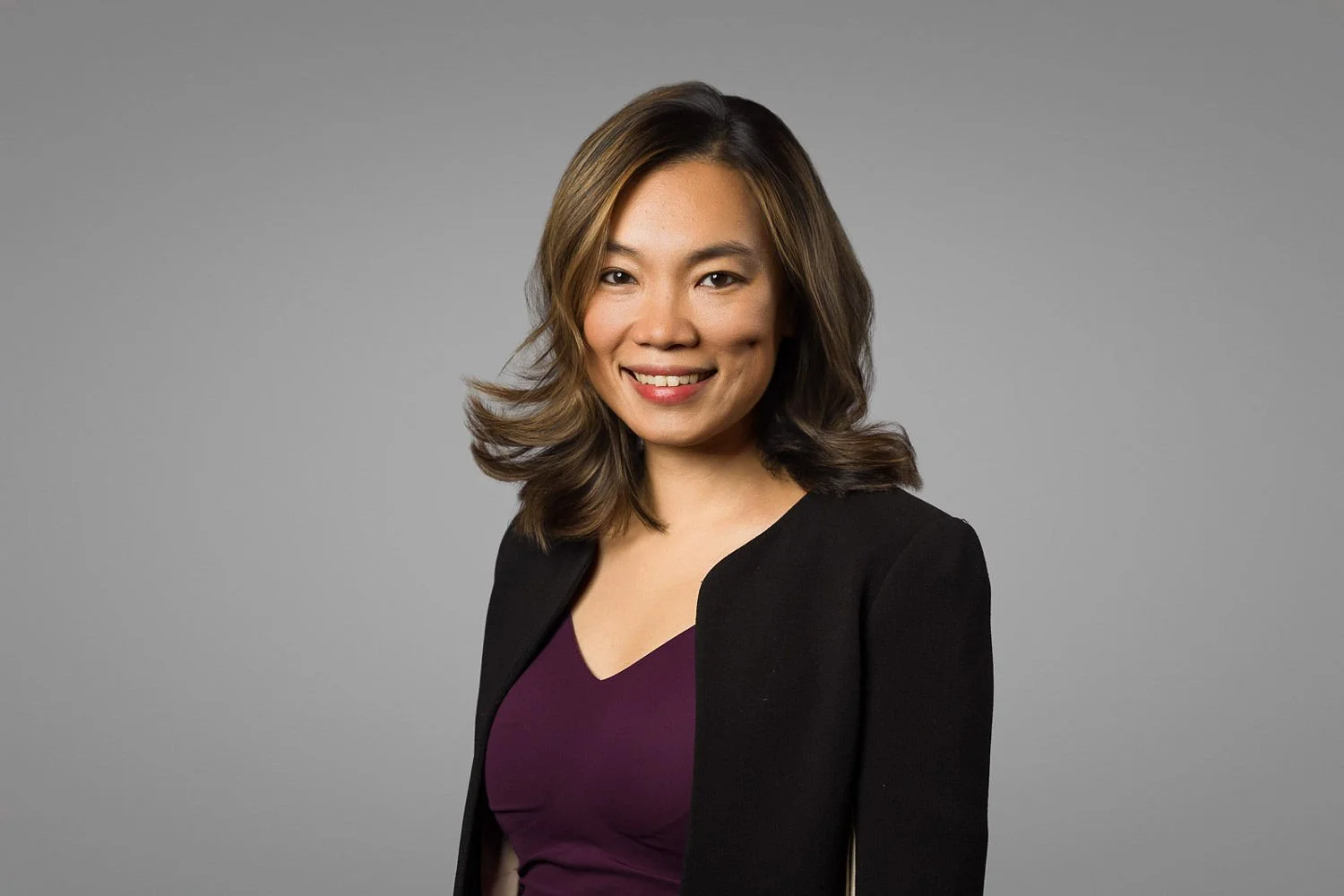 A professional woman with shoulder-length wavy brown hair, smiling, wearing a black blazer over a purple top, standing against a gray background.