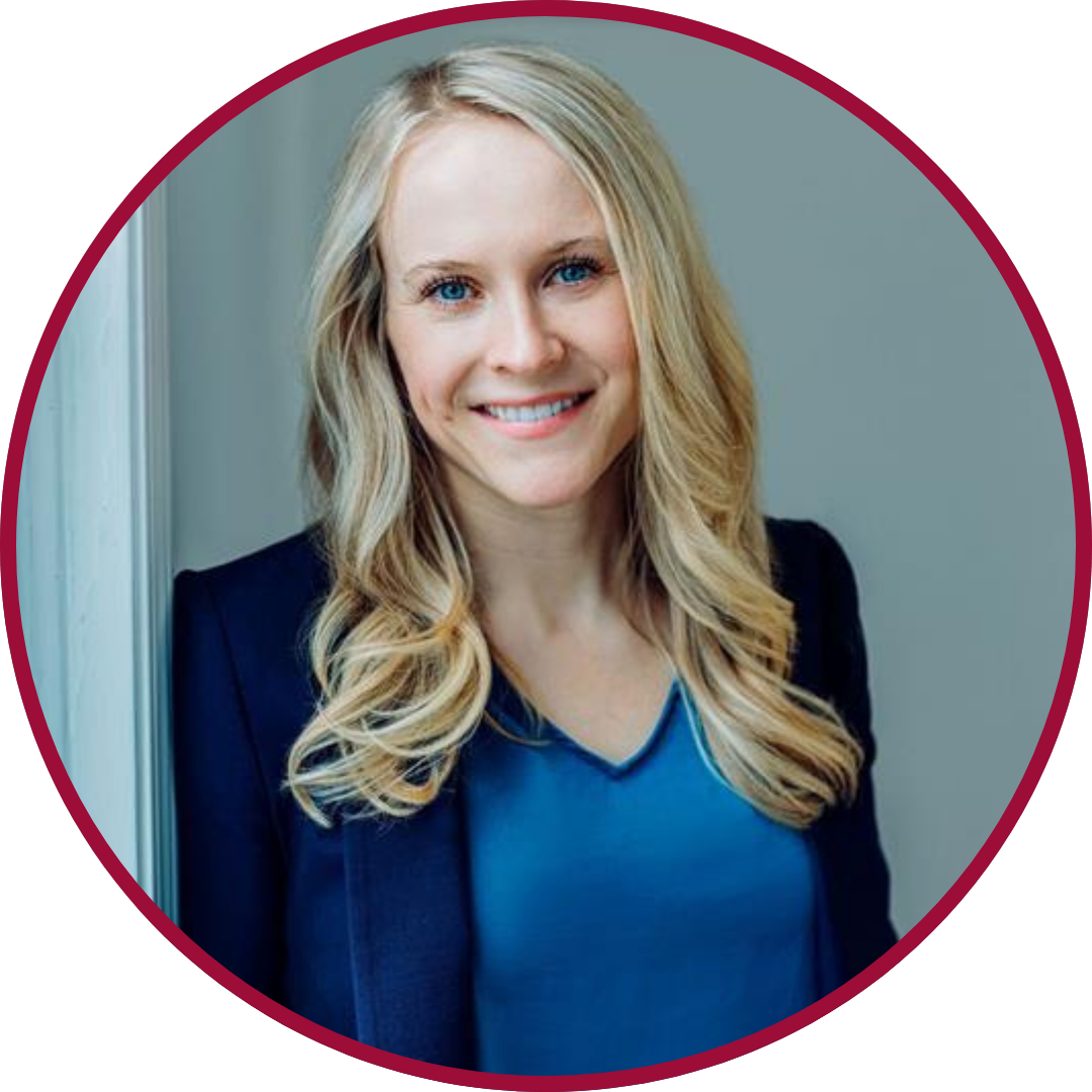 A woman with long blonde hair, blue eyes, and a bright smile, wearing a blue blouse and a dark blazer, standing near a window with a neutral wall background.