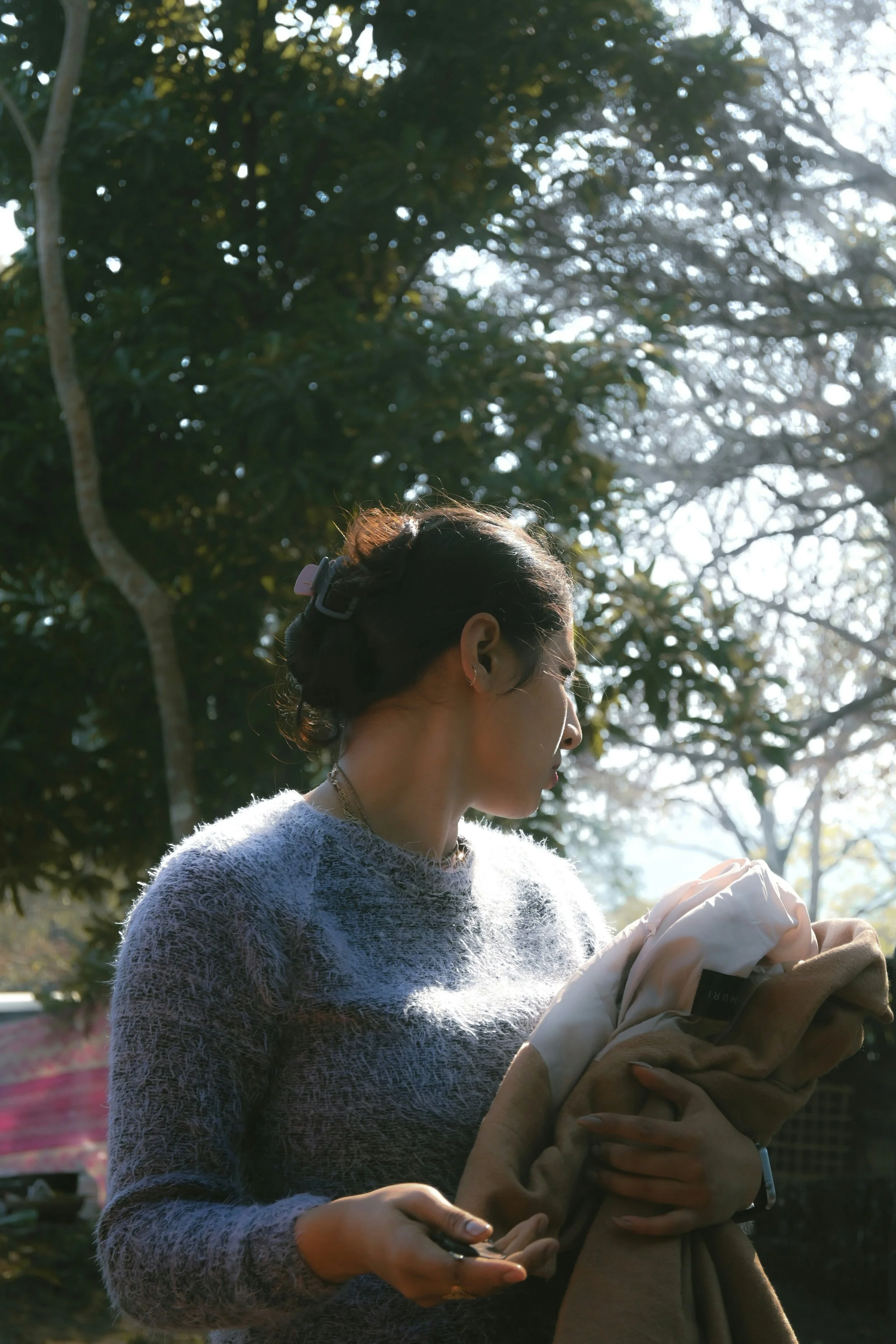 A woman standing outdoors, holding a bundle of clothes, with sunlight filtering through trees in the background.