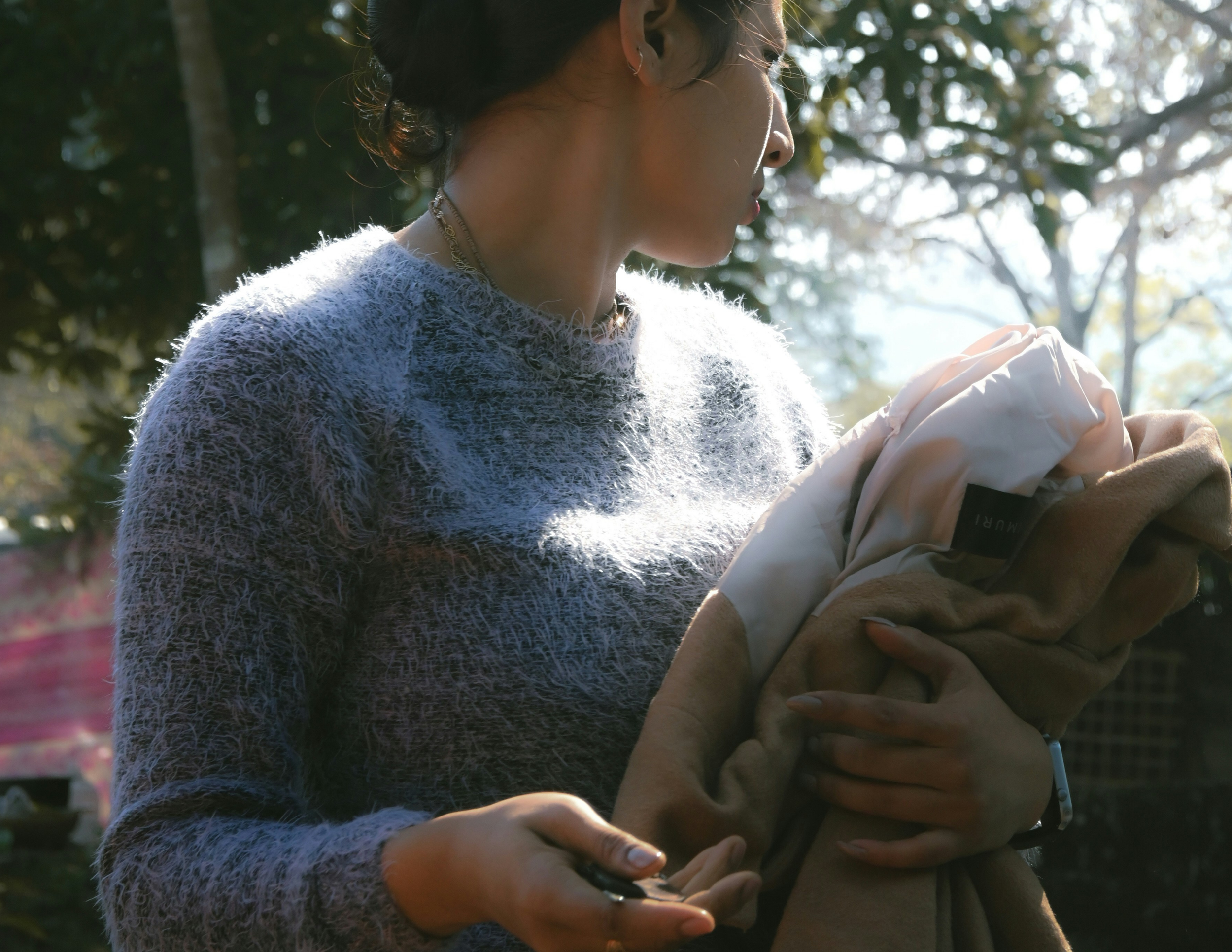 A woman holding a baby wrapped in a blanket outdoors in a forested area with sunlight filtering through trees.