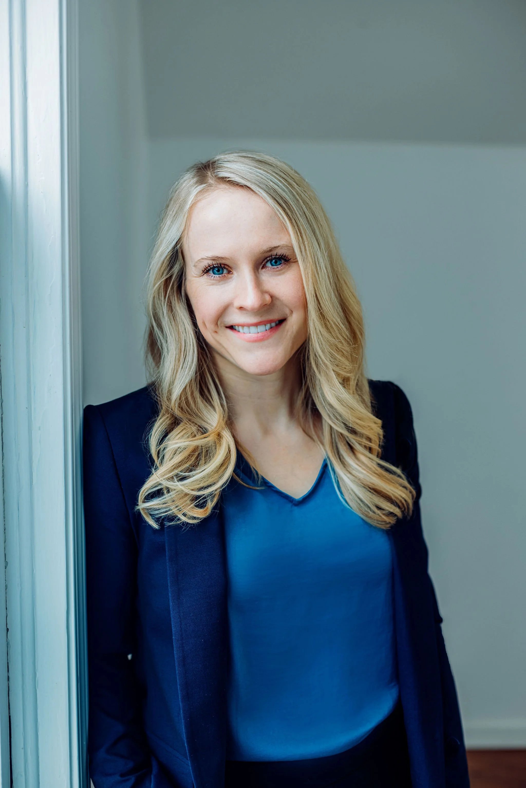 A woman with blonde hair, blue eyes, and a bright smile, wearing a blue blouse and a navy blazer, standing indoors near a doorway.