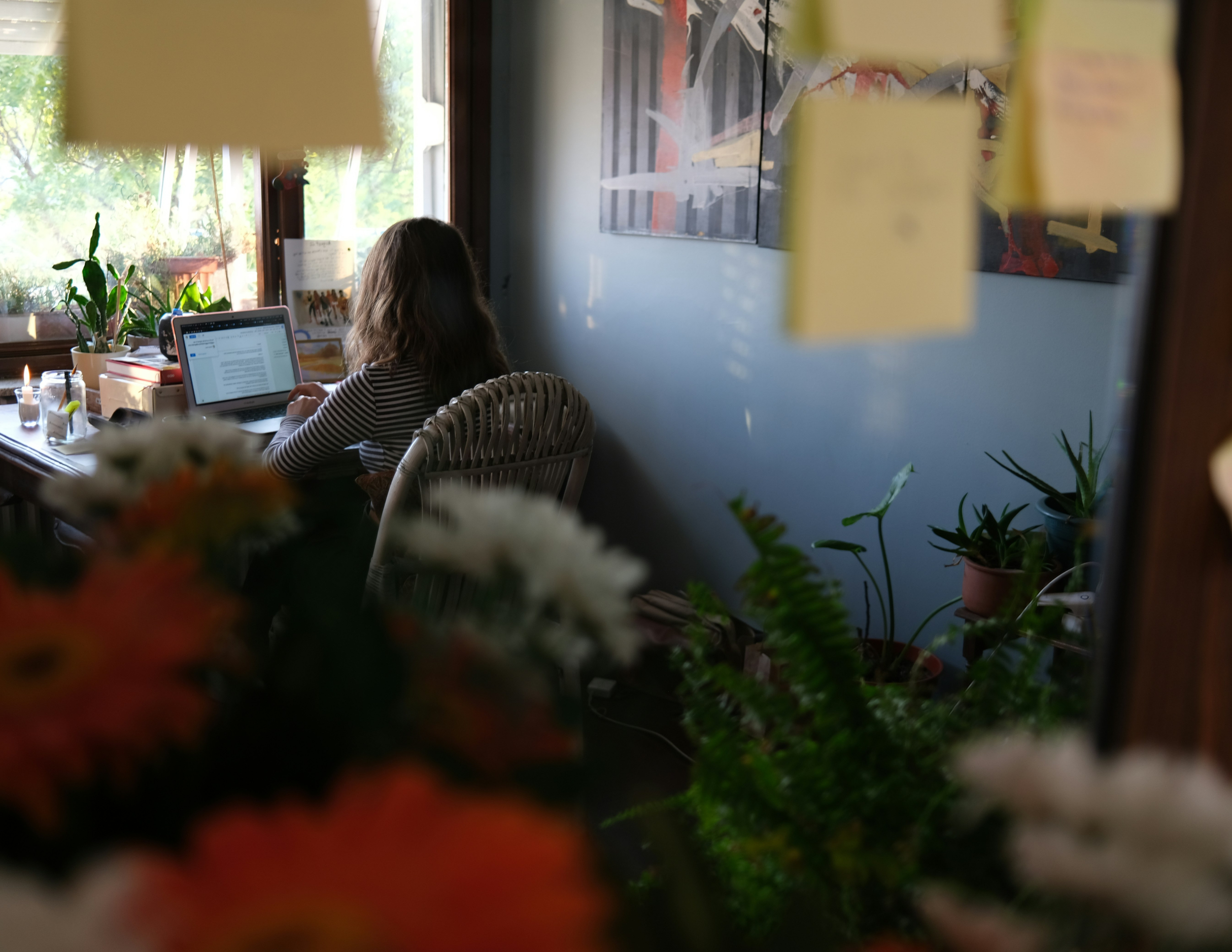 A person sitting at a desk with a laptop, looking out a window. The desk is decorated with plants, candles, and books. The scene is cozy with natural light coming through the window.