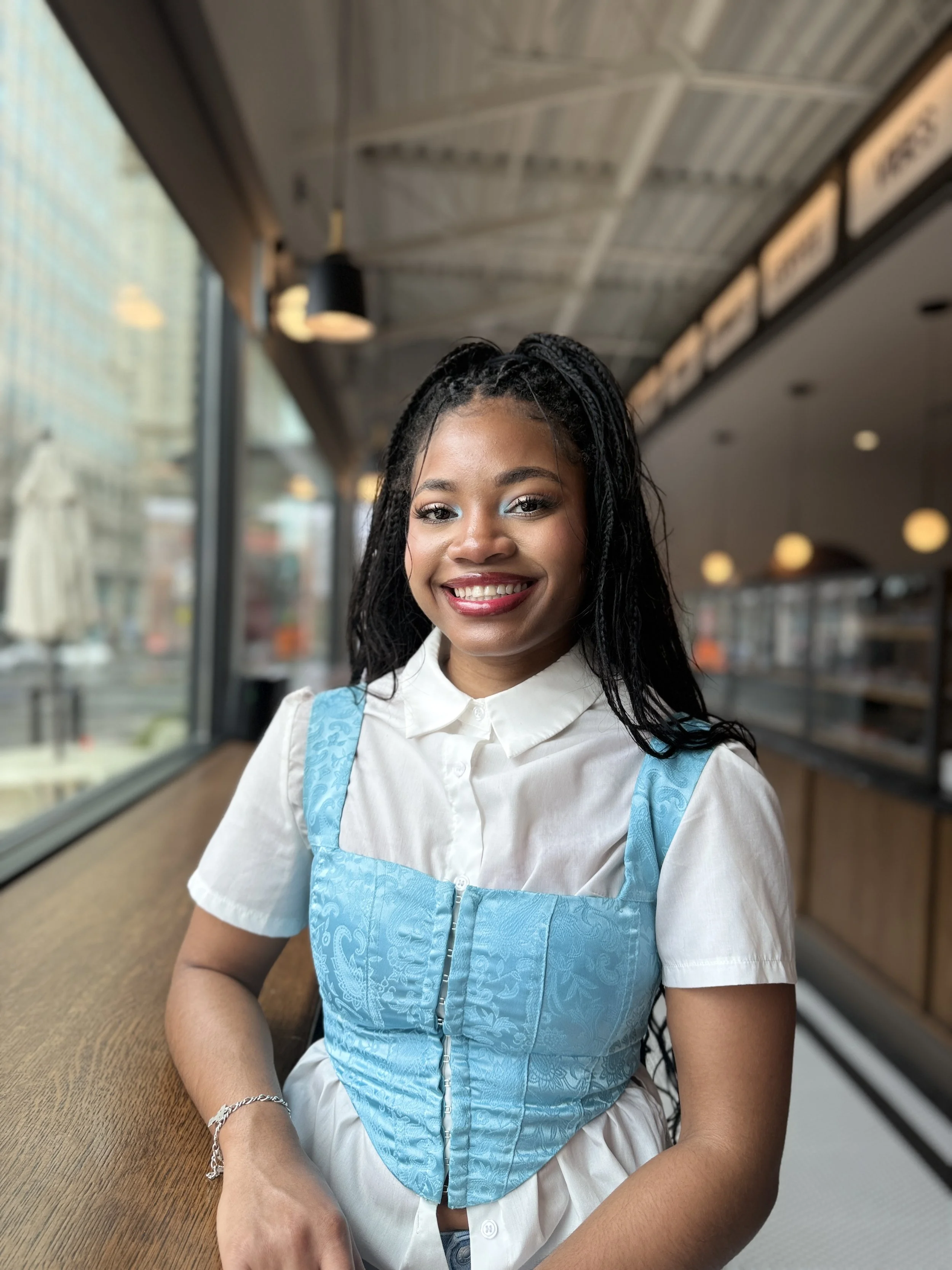 Young woman with long black hair braided on top, smiling, wearing a white shirt and a blue vest, sitting at a cafe with large windows and indoor lighting.