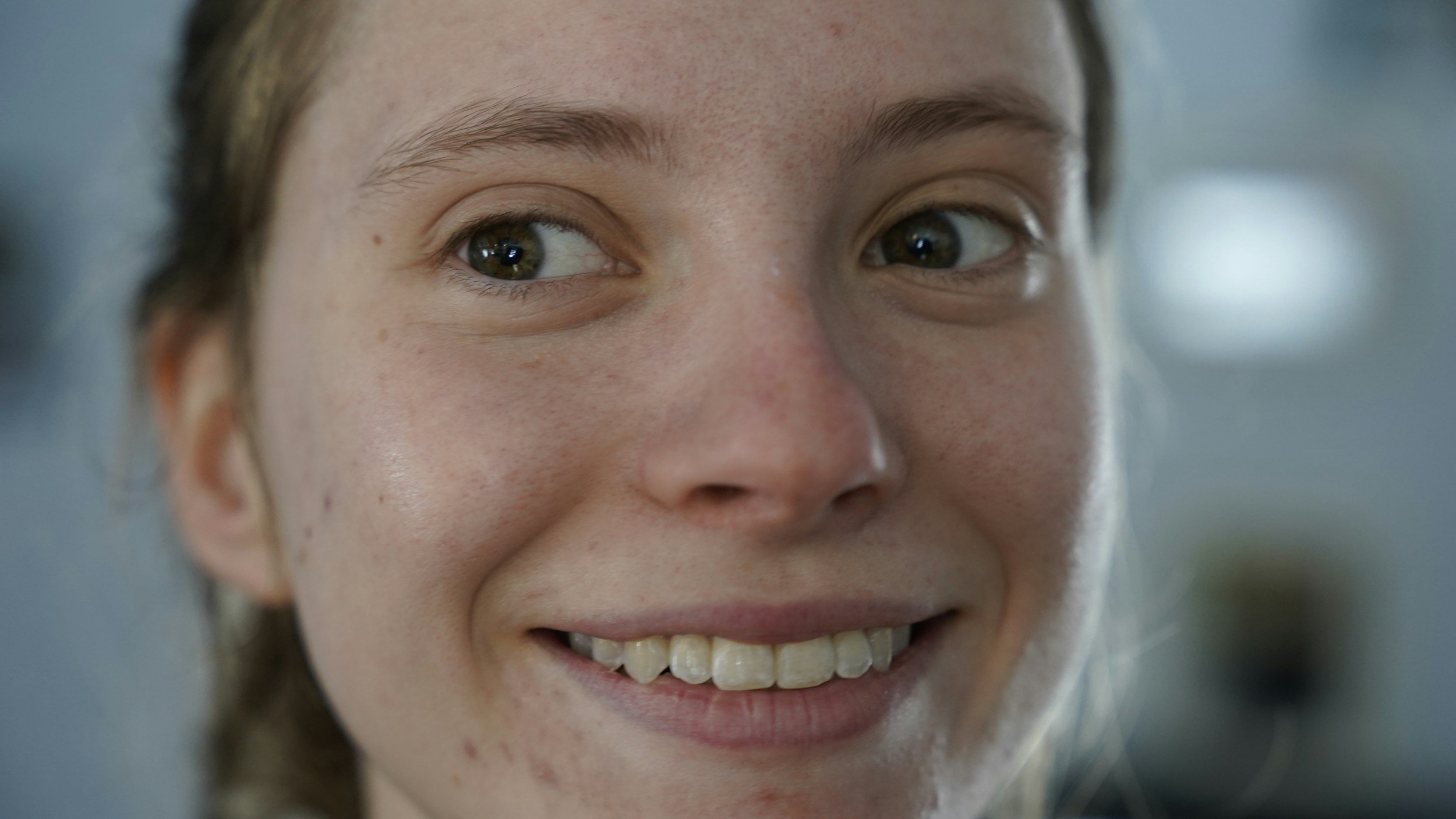 Close-up of a young woman's face with light skin, brown eyes, and smiling mouth showing teeth.