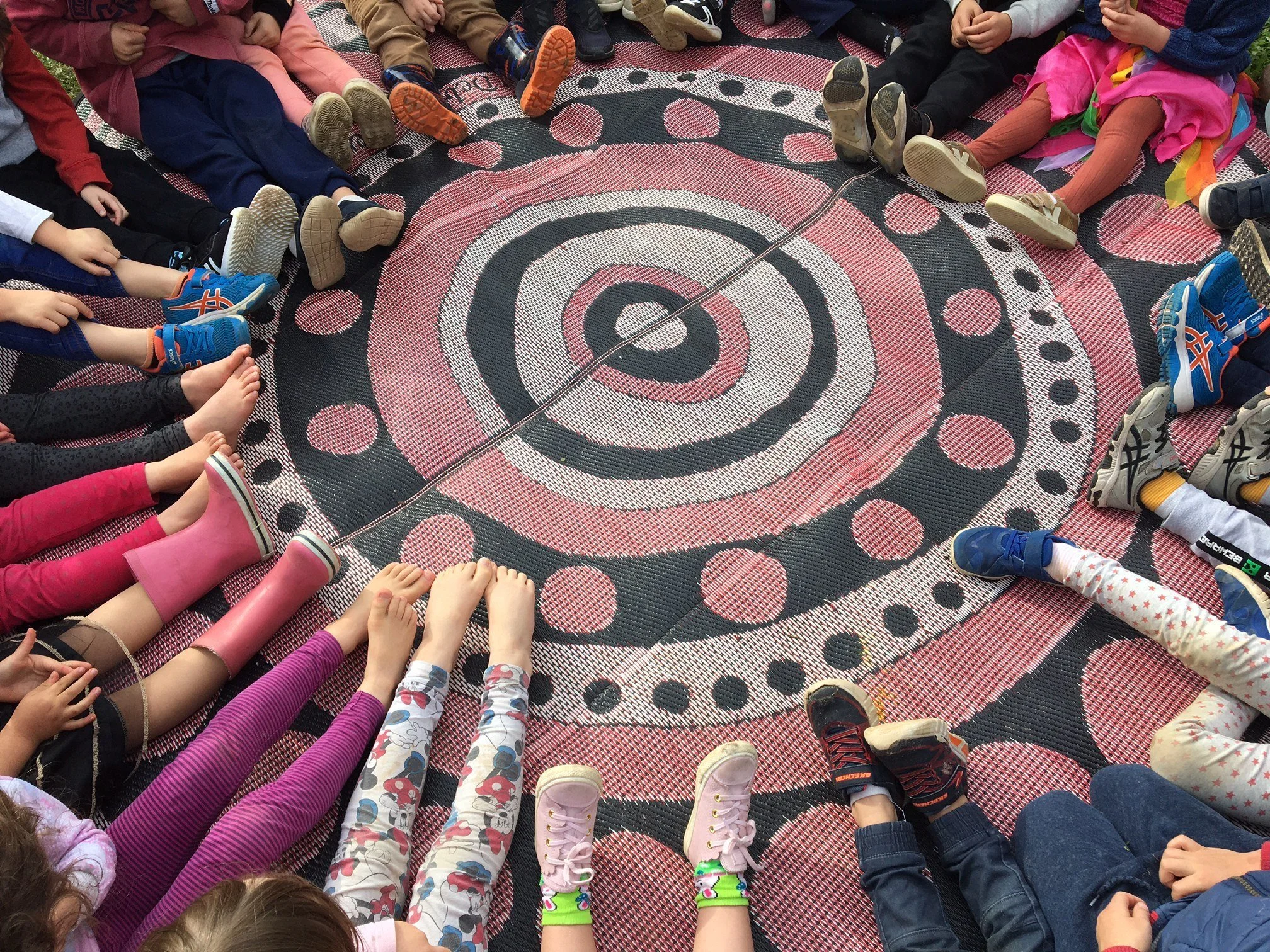 Children sitting in a circle with their legs extended outward on a large circular rug with concentric circle and dot patterns.