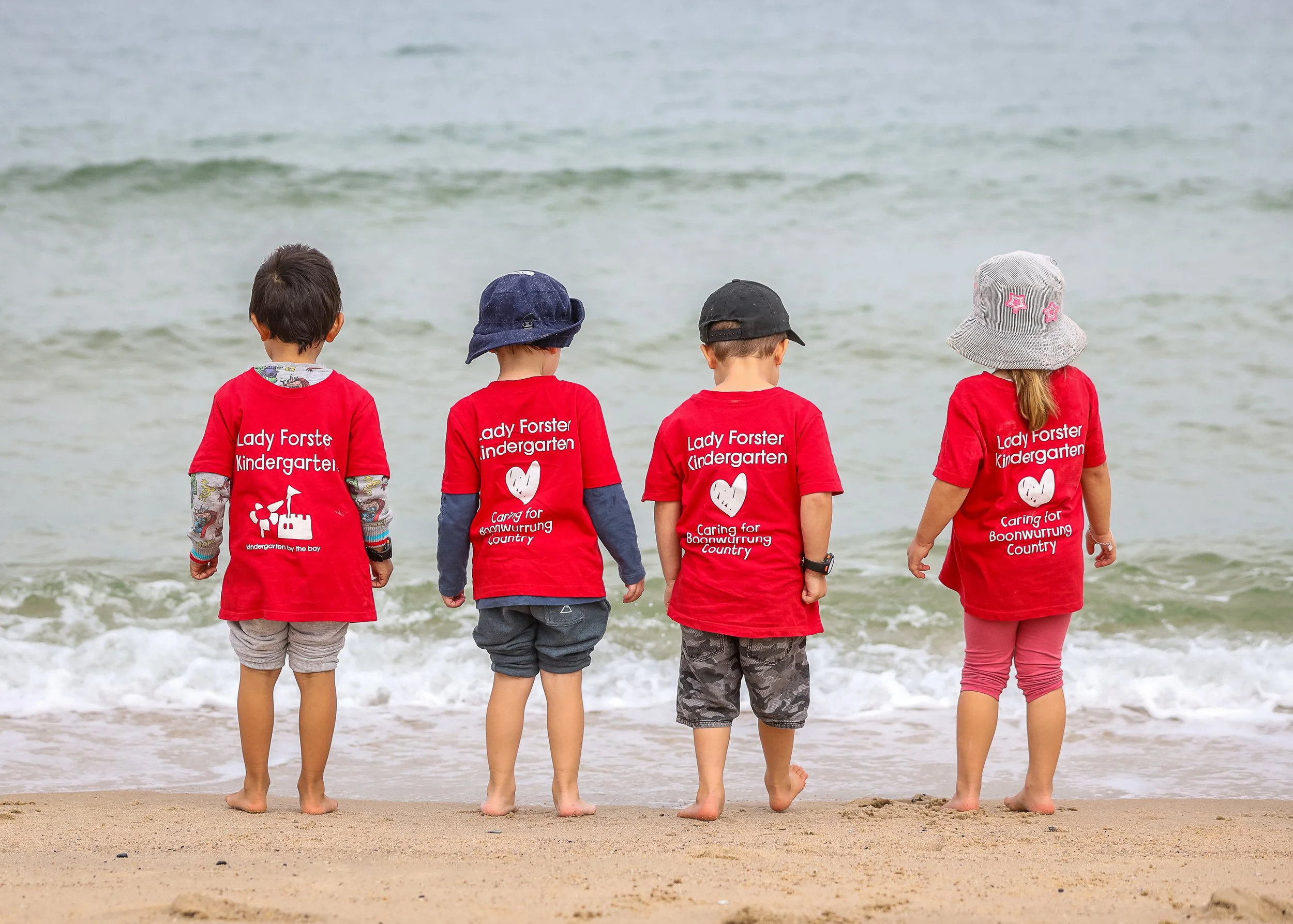 Four young children standing on the beach facing the ocean, wearing red T-shirts that read 'Lady Forster Kindergarten' with a heart and slogan, some wearing hats, with waves in the background.