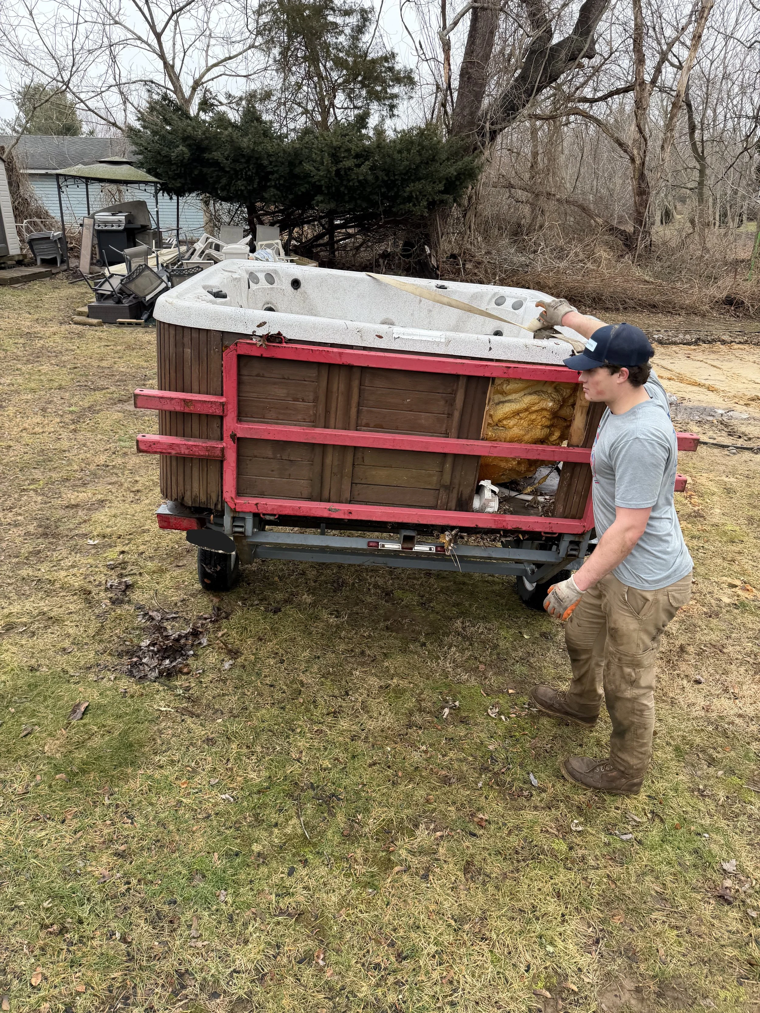 A person standing outdoors next to a hot tub on a metal platform, with insulation exposed on the side. The person is wearing gloves, a gray t-shirt, tan pants, and a navy baseball cap, and appears to be working on the hot tub. The background shows a 