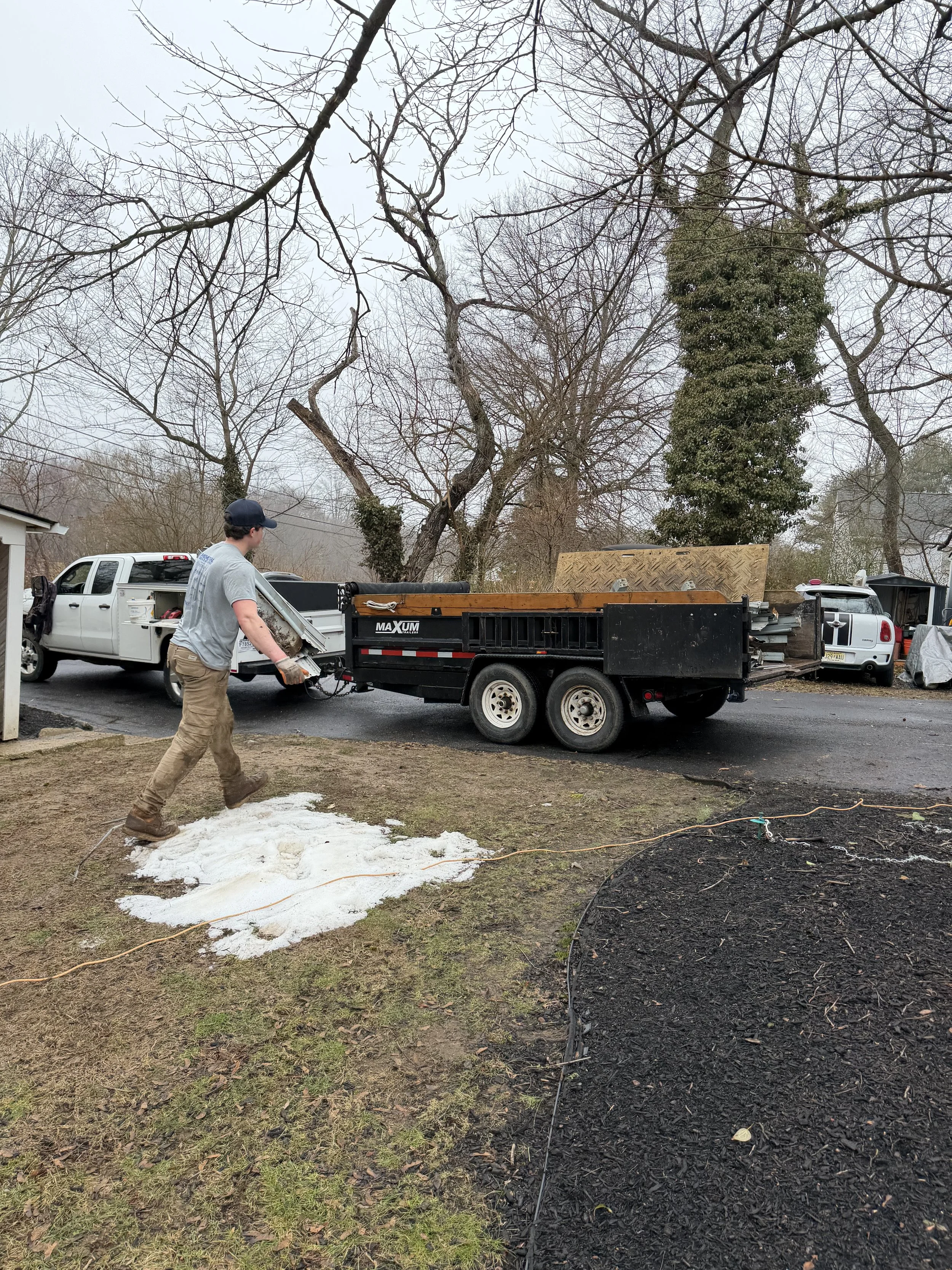 A man walking on grass in a yard next to a patch of snow, while a utility trailer connected to a white truck is parked on a driveway. The trailer holds various equipment and materials, with trees and other vehicles in the background.