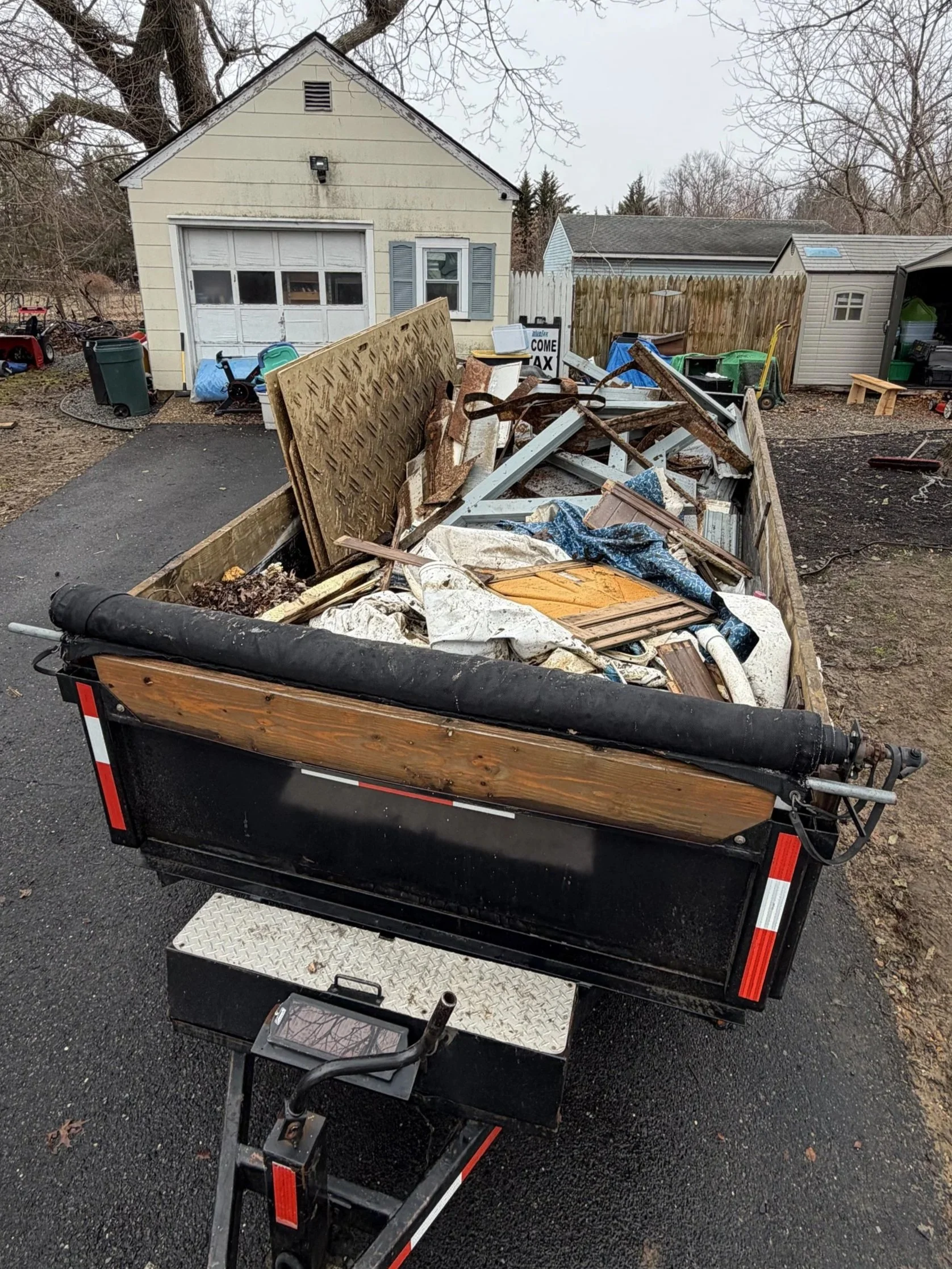 A trailer filled with construction debris, including wood panels, metal, and other materials, parked on a paved driveway in a residential backyard.