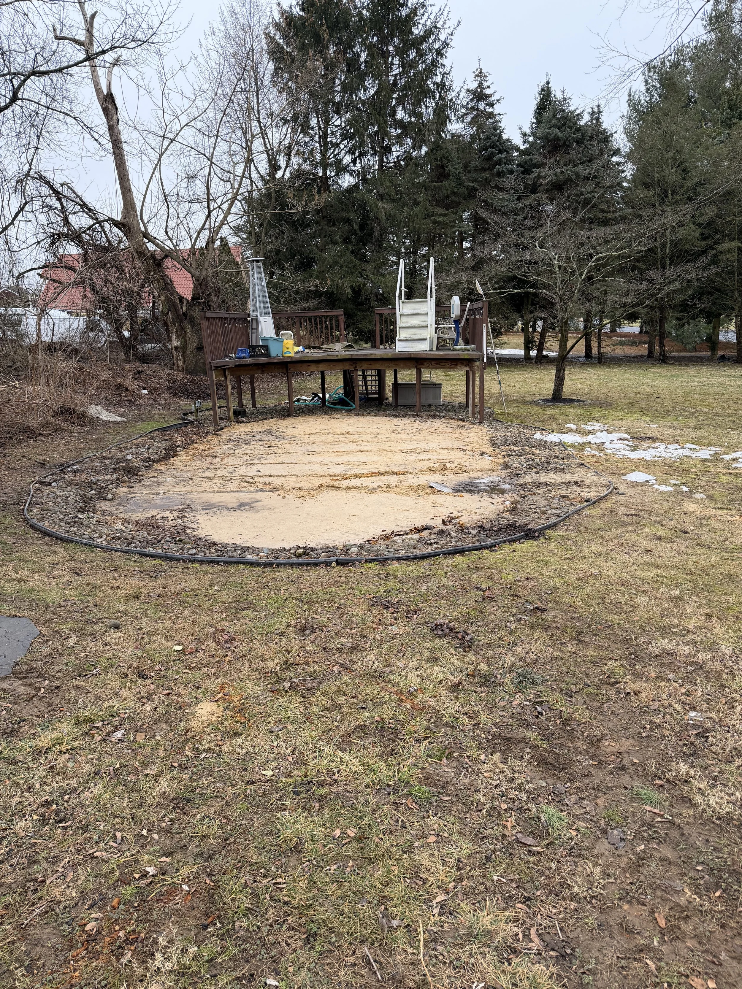 Backyard scene with a wooden deck and stairs leading up to it, surrounded by trees and a grassy yard. The ground in front has been prepared for a new surface, with some sand spread out. There are some patches of snow on the grass, and equipment is vi