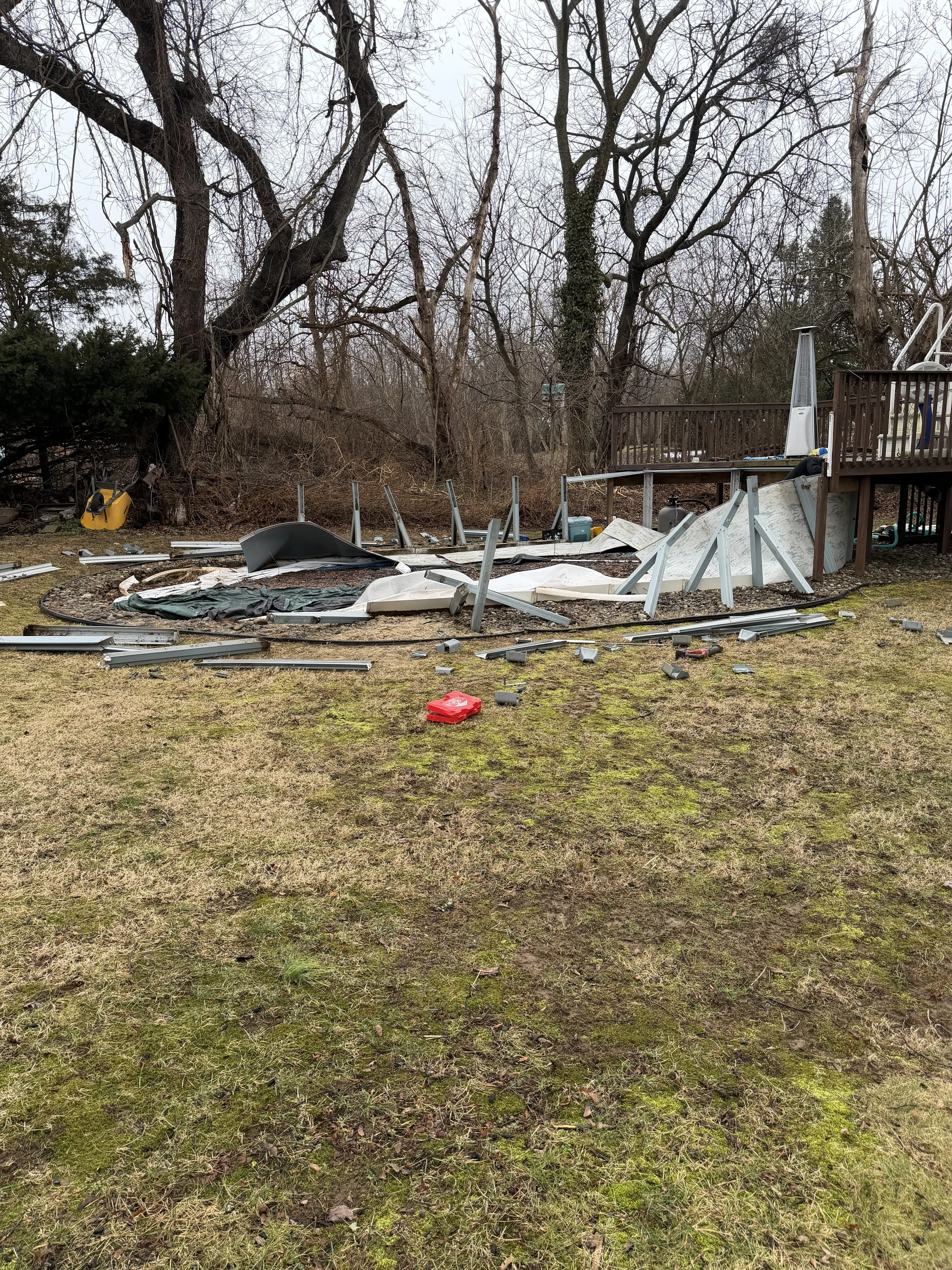 Yard with collapsed and broken backyard swimming pool and dismantled metal fence panels, debris scattered across the ground, with leafless trees in the background.