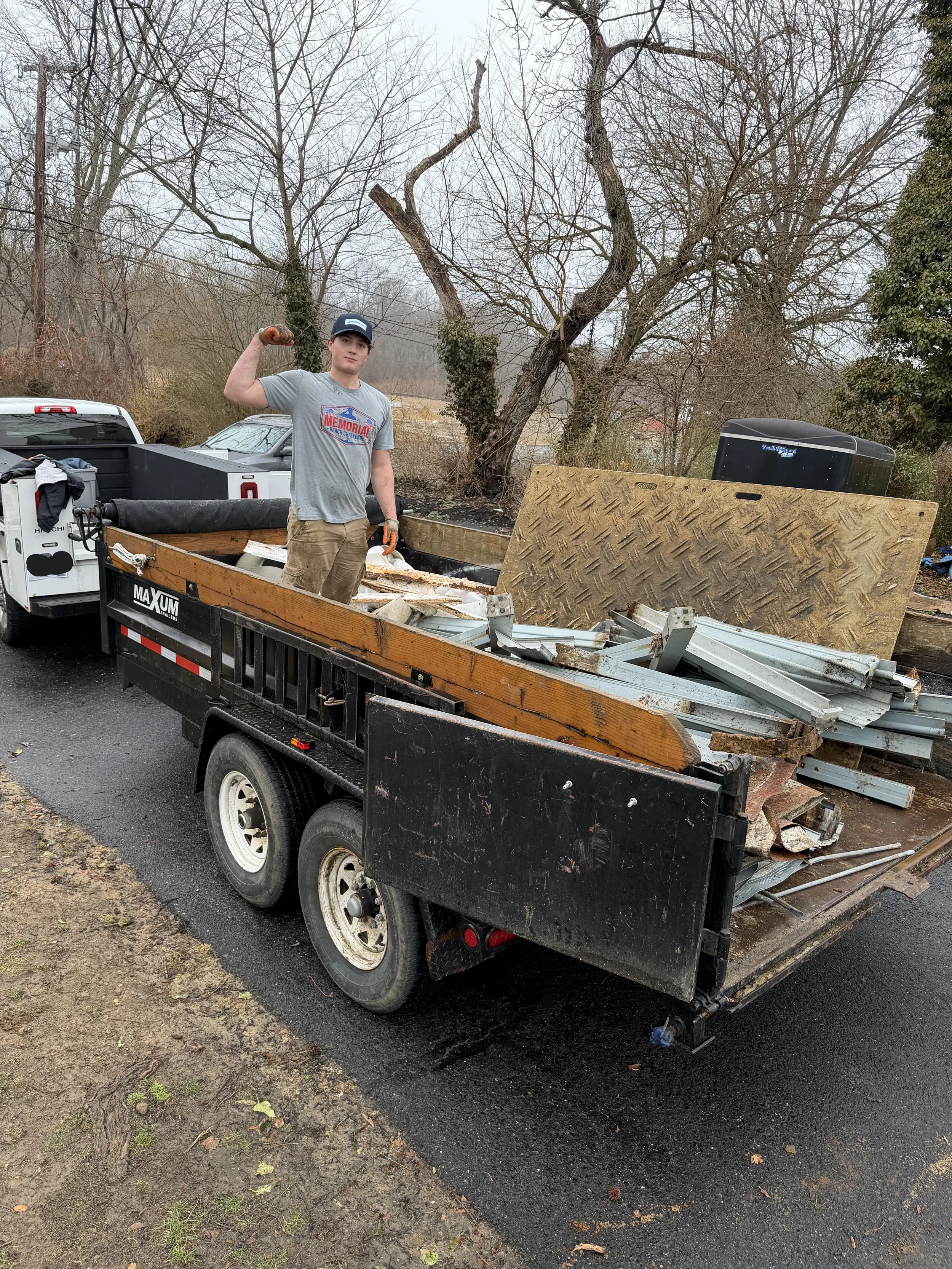 A young man standing on a flatbed trailer filled with construction debris, including metal, wood, and other materials, holding a small rock or chunk of debris in one hand and wearing a baseball cap and gloves, with a leafless tree and a black trailer