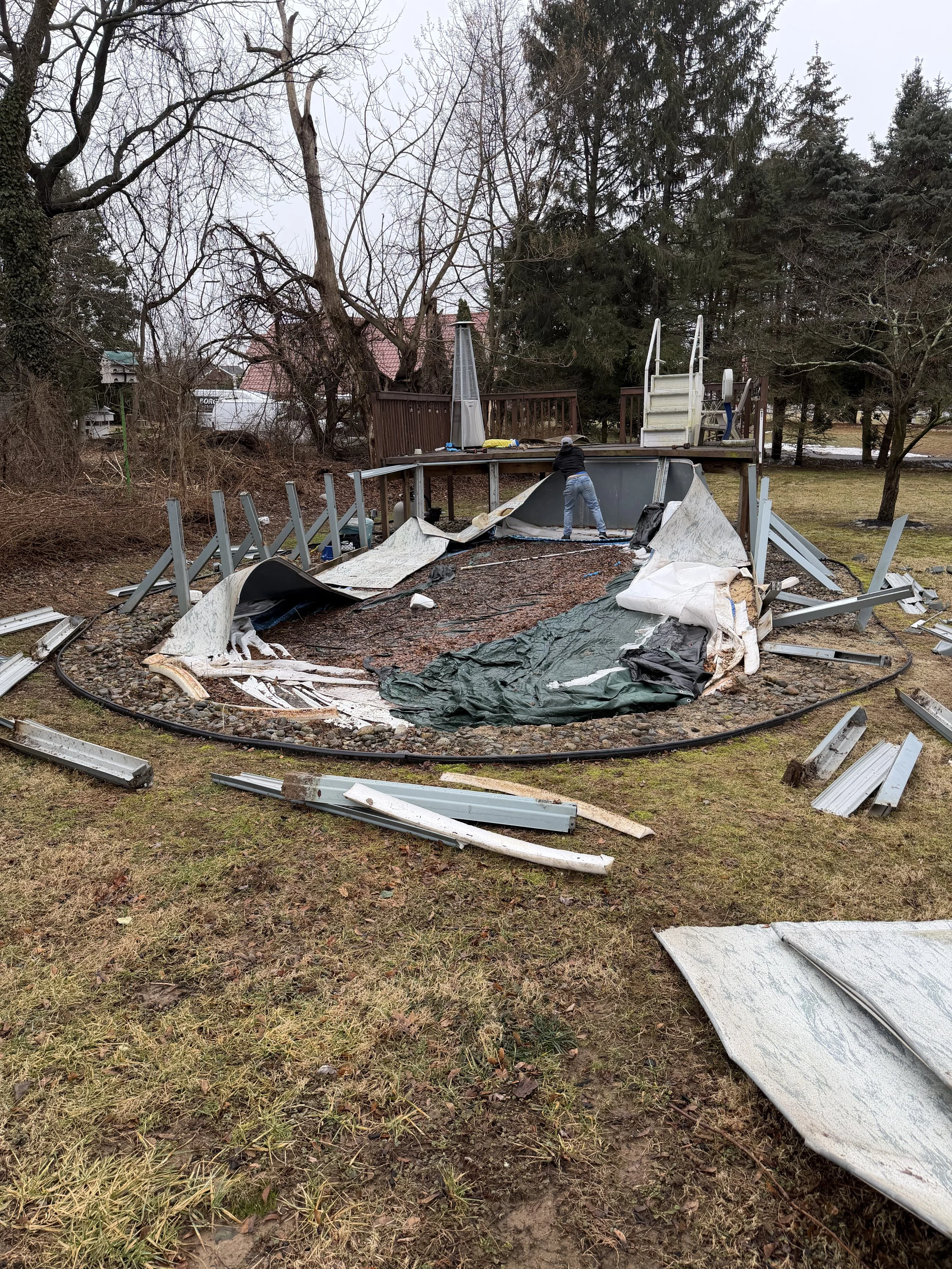 Collapsed outdoor pool area with debris, roof panels, and a person working on cleanup on cloudy day.