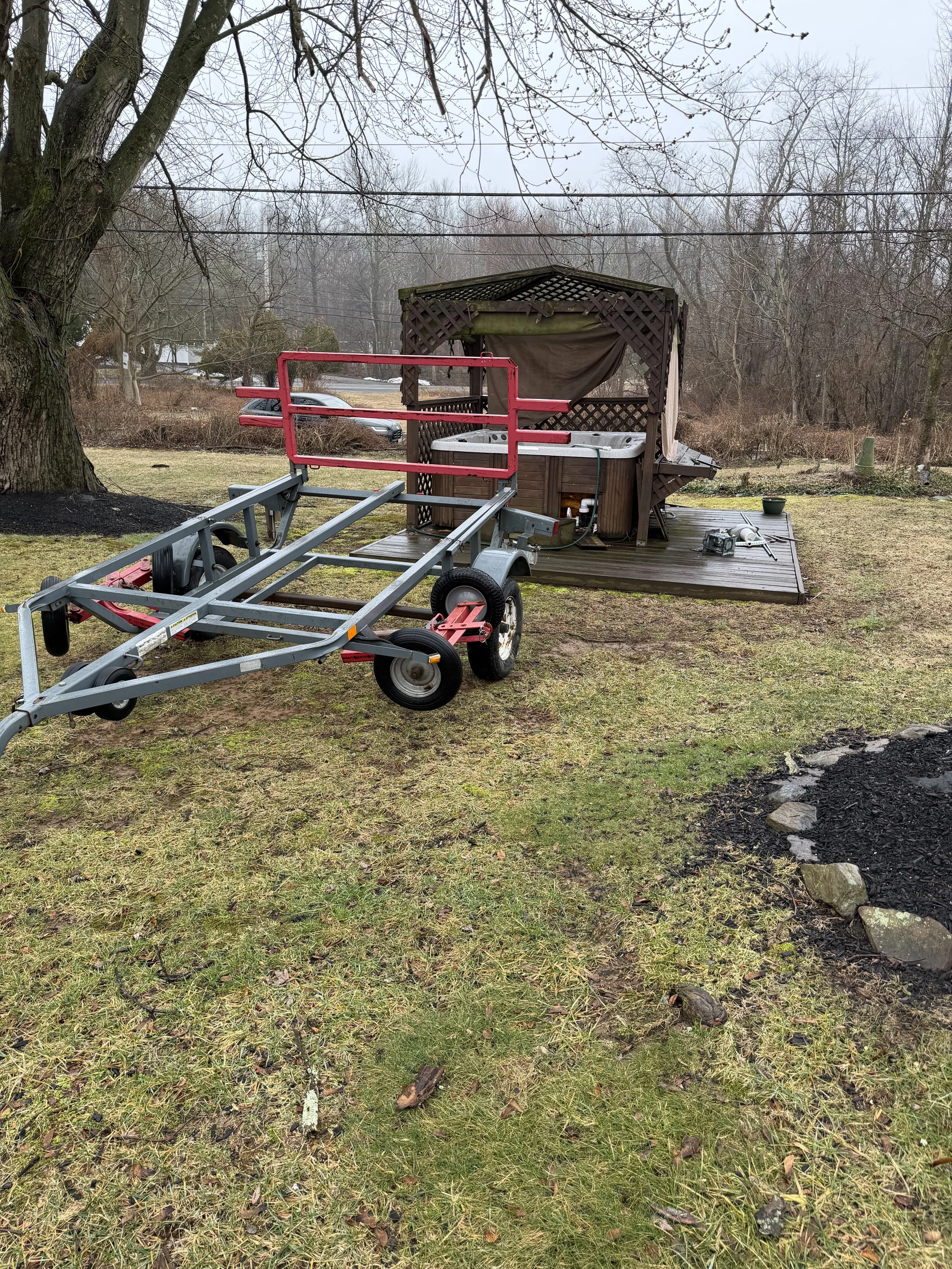 A hot tub on a wooden deck with a canopy, with a boat trailer parked in front of it on patchy grass yard, overcast sky, leafless trees, and a tree on the left side.