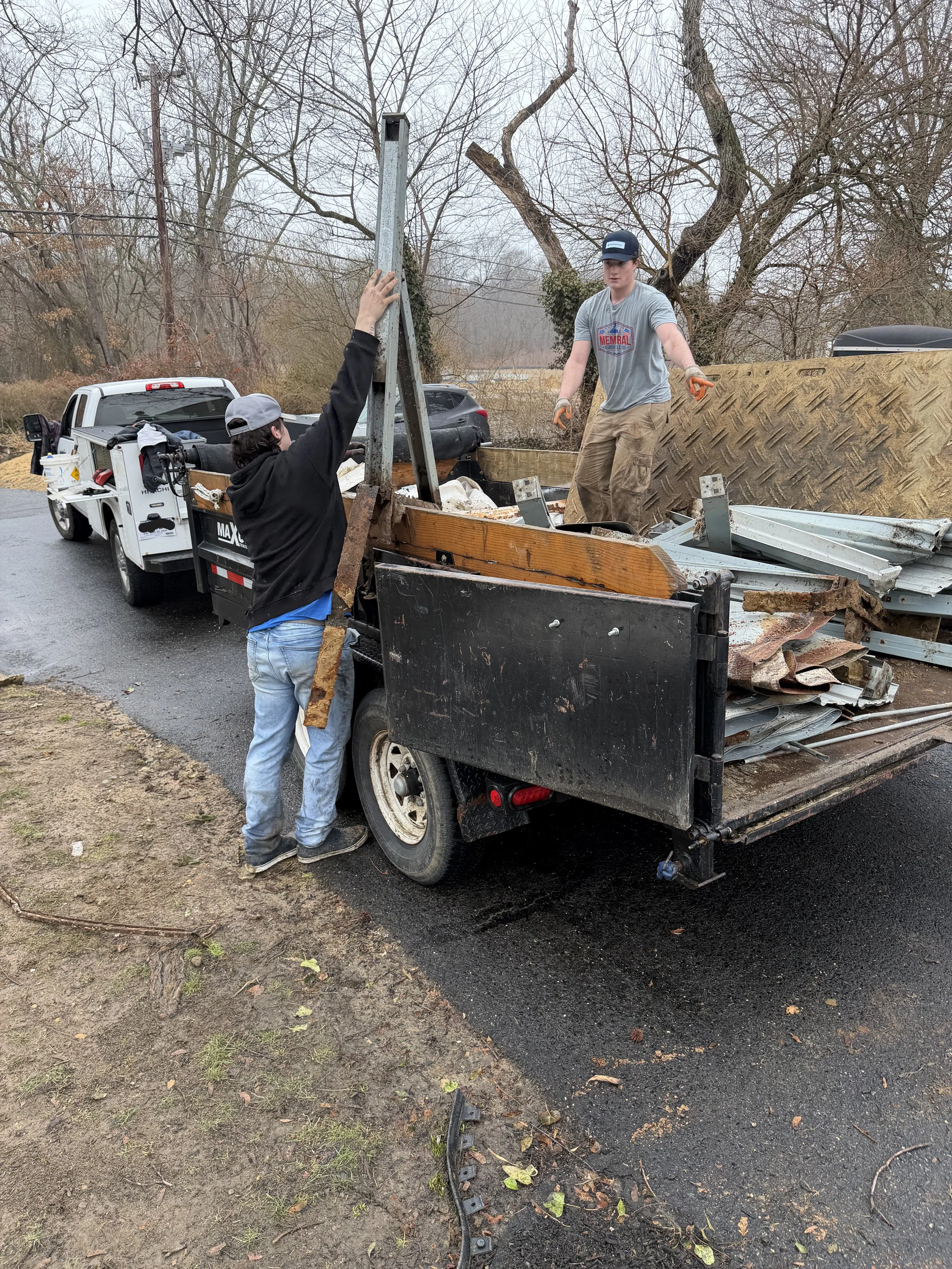 Two men are loading debris onto a flatbed truck, with one man standing beside the truck and the other standing on the truck's bed. They are working outdoors near a road lined with leafless trees, in a cloudy, overcast setting.