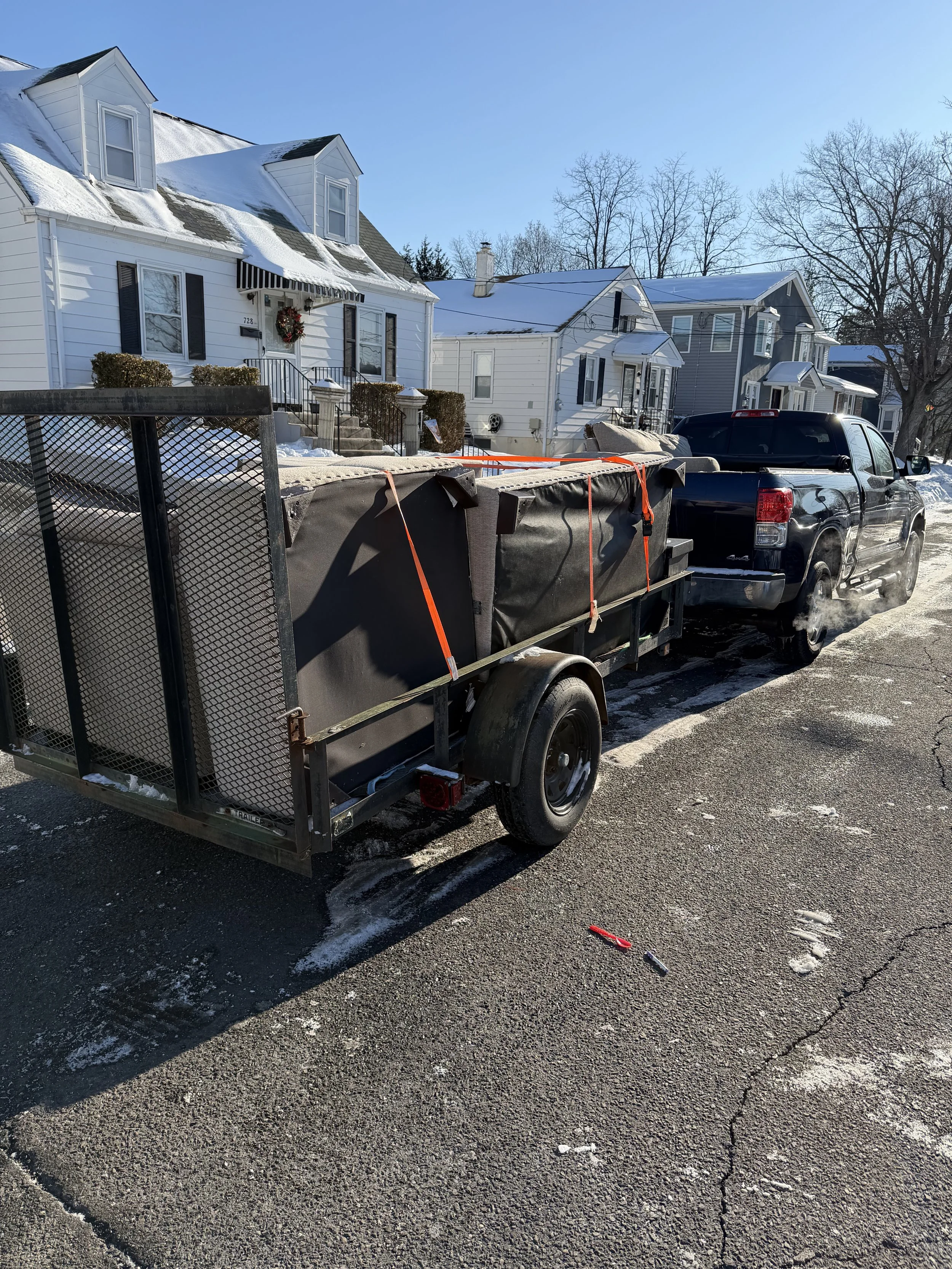 Black pickup truck towing a black trailer with snow removal equipment, parked on a snow-dusted street in a residential neighborhood with snow-covered houses and leafless trees.
