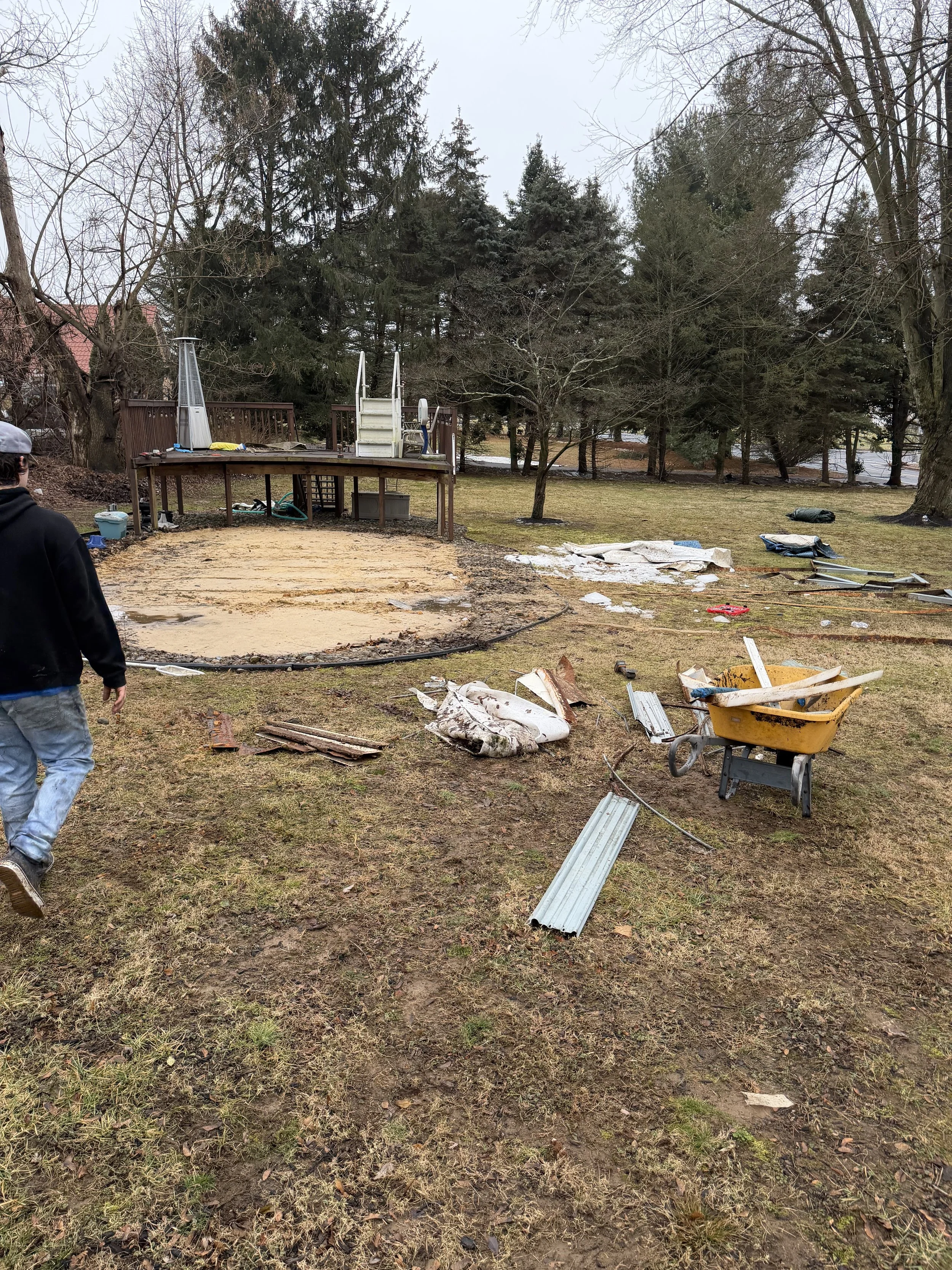 Yard with debris, broken materials, and a swing set removed, indicating recent cleaning or renovation. A person is walking away on the left side, and trees are in the background.