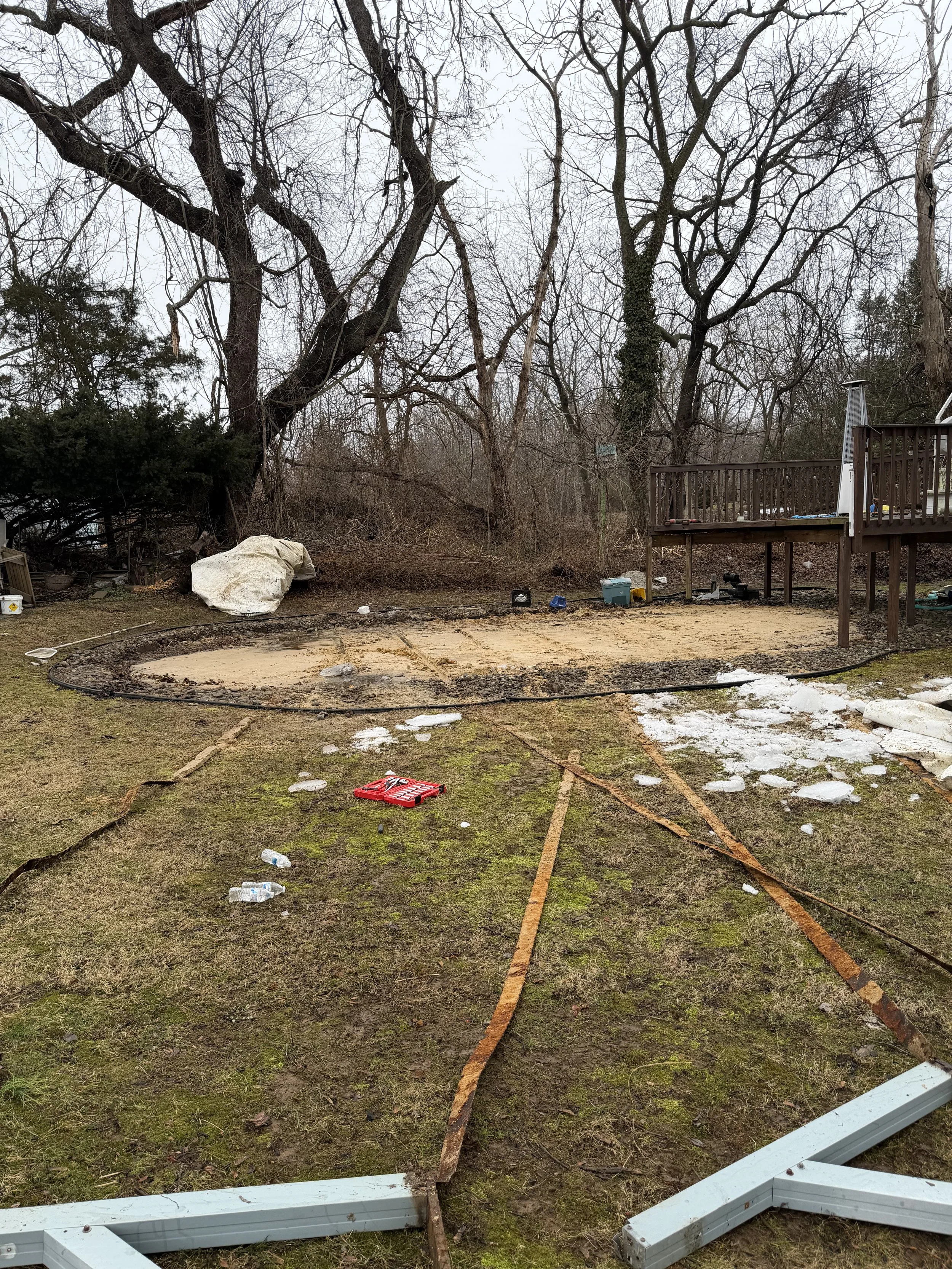 Backyard with a partially dismantled deck, scattered debris, tools, and trash, with leafless trees in the background on a cloudy day.