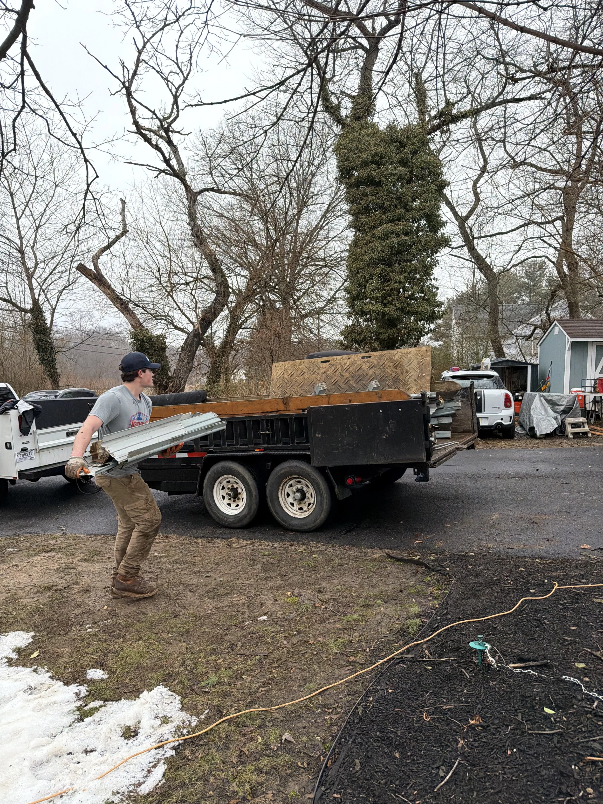 A man unloading metal roofing panels from a flatbed trailer attached to a white pickup truck in an outdoor area with leafless trees and some snow on the ground.