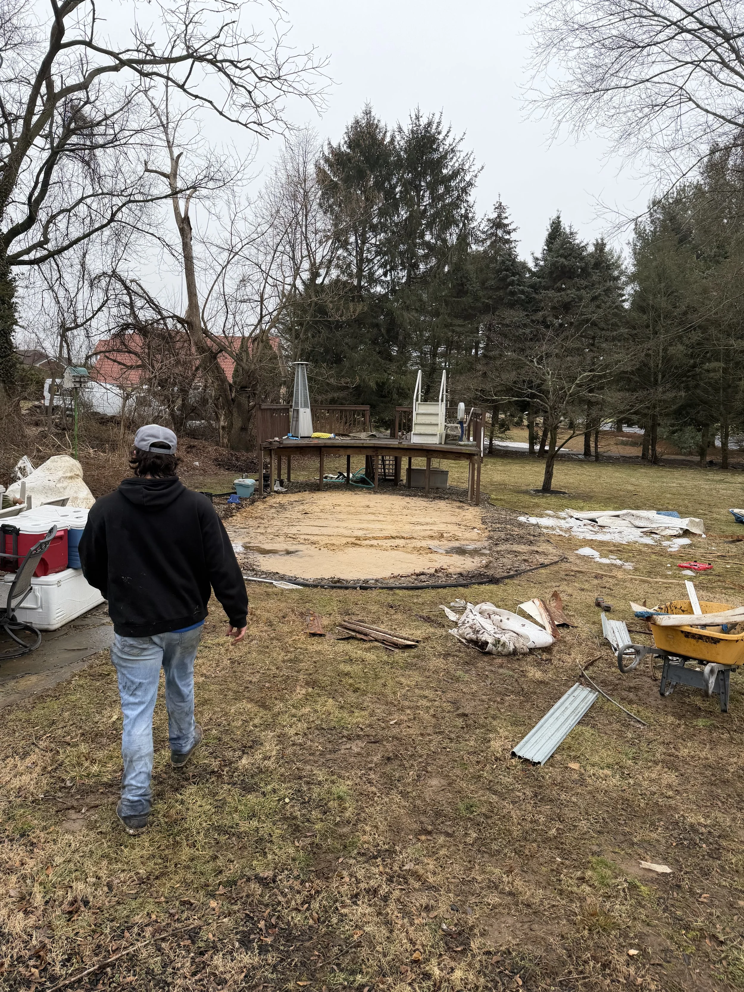 A backyard with a large deck and stairs, surrounded by trees, with debris and scattered items including a wheelbarrow, a cooler, and trash, on a cloudy day.