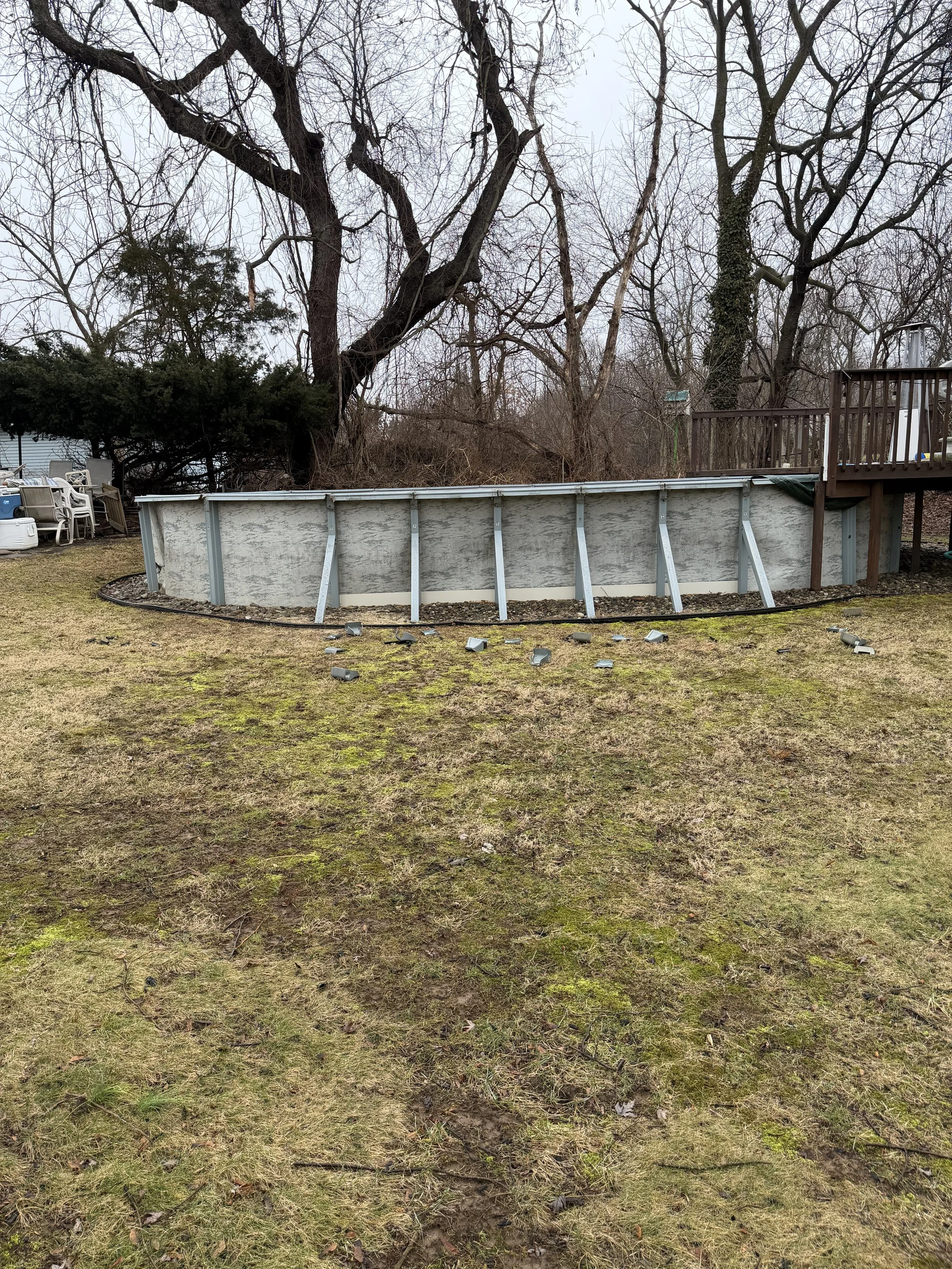 A backyard with a above ground swimming pool with the liner removed, leaving the metal frame exposed. The ground nearby is patchy with grass and moss, scattered with fallen debris and small objects. In the background, leafless trees and a wooden deck