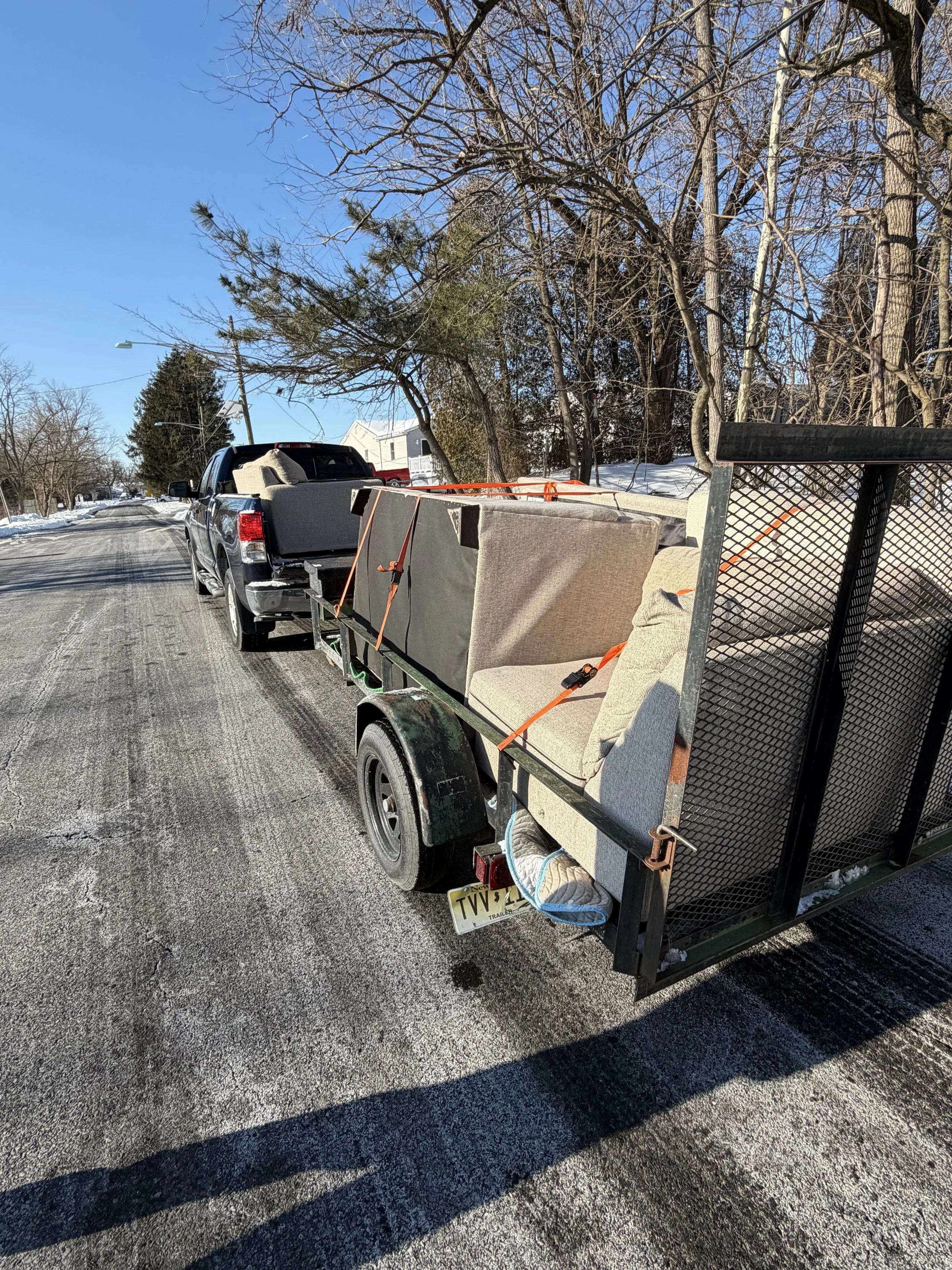 A pickup truck towing a trailer with furniture, including couches and an armchair, secured with orange straps on a cold, snowy street with leafless trees.