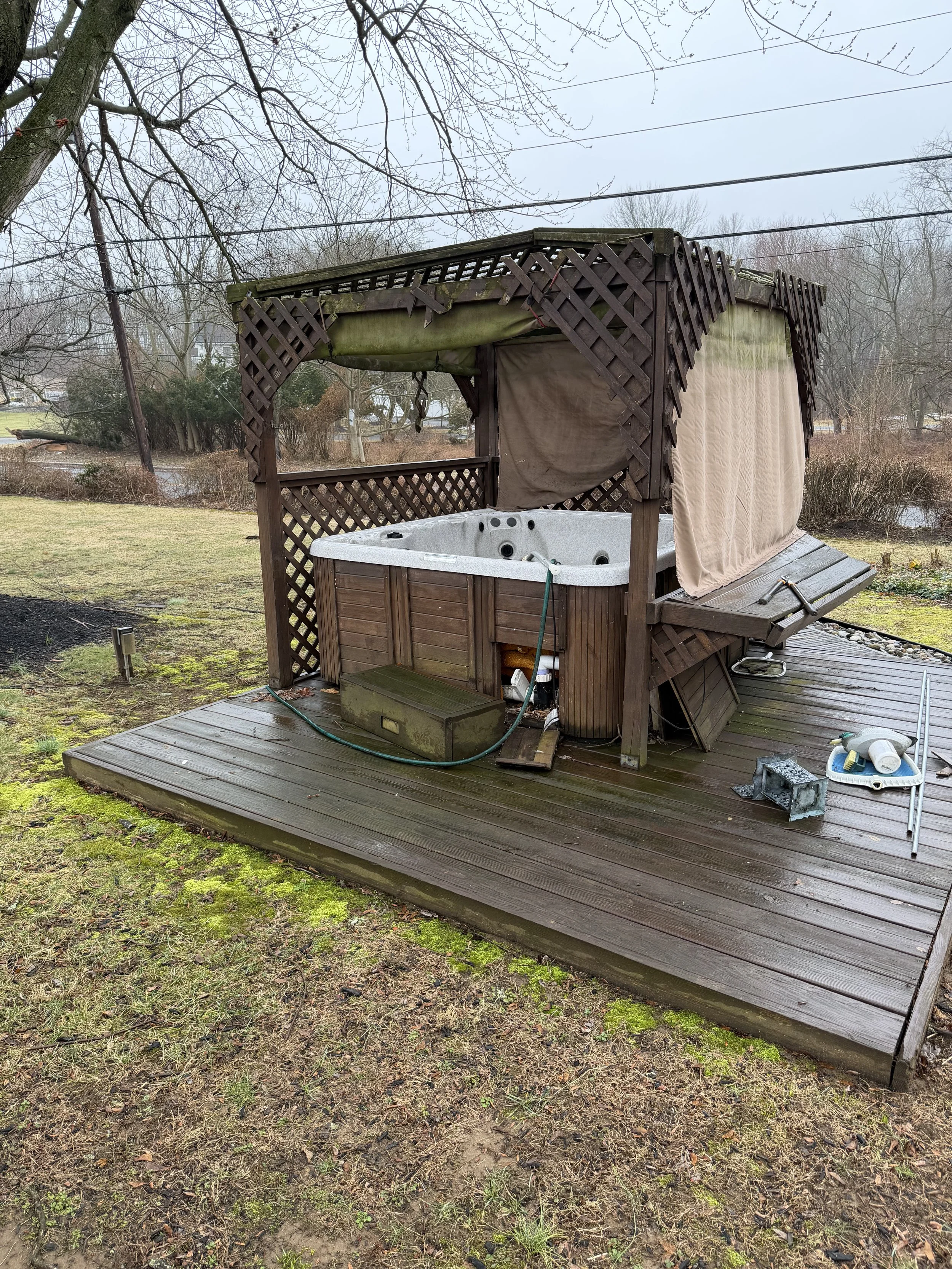 Outdoor hot tub on a wooden deck with green moss and debris, surrounded by a grassy yard with trees and a pond in the background.