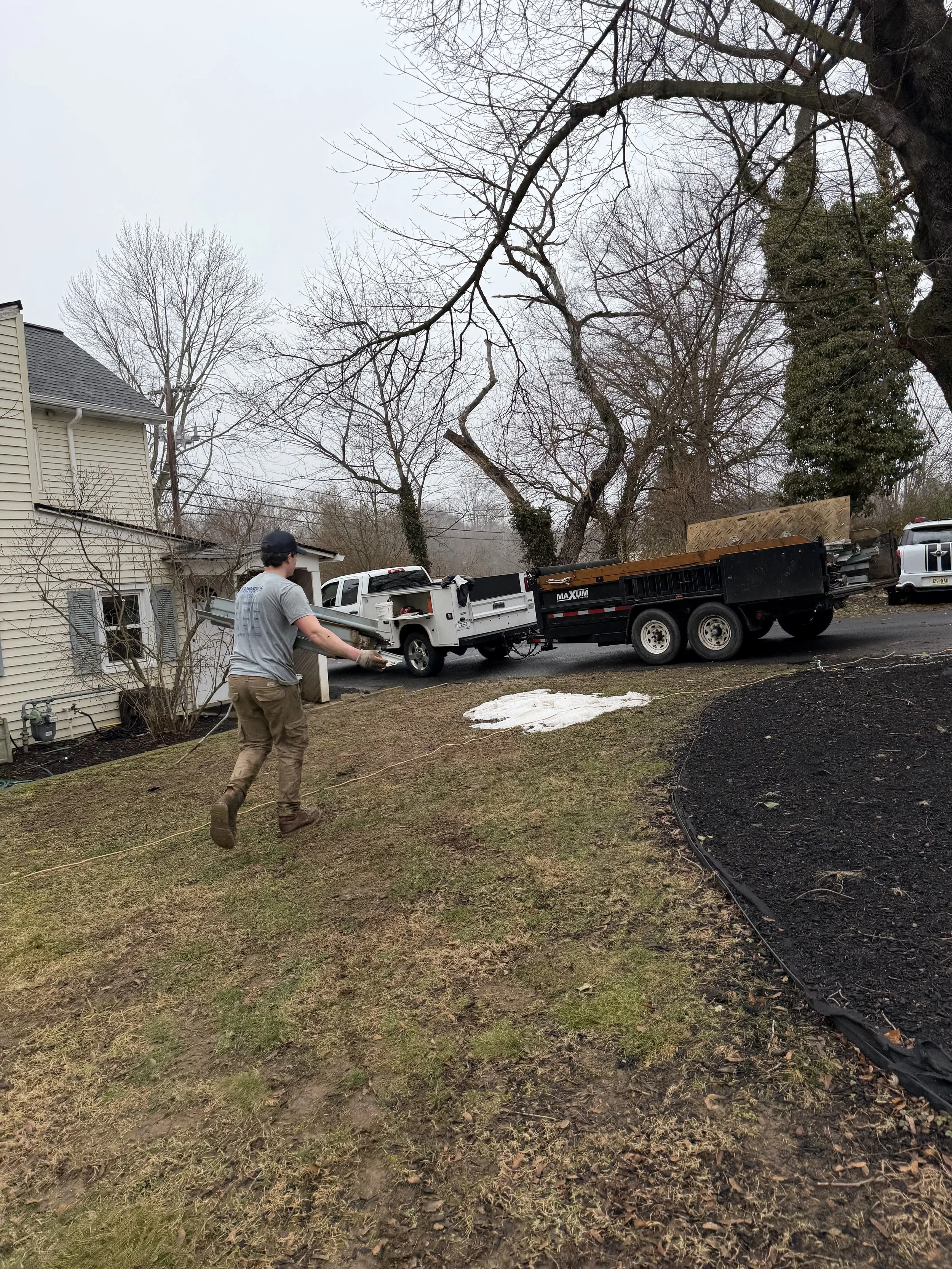 A man working outside on a cloudy day near a house, pulling a rope or hose, with trucks and construction equipment in the driveway, and leafless trees in the background.