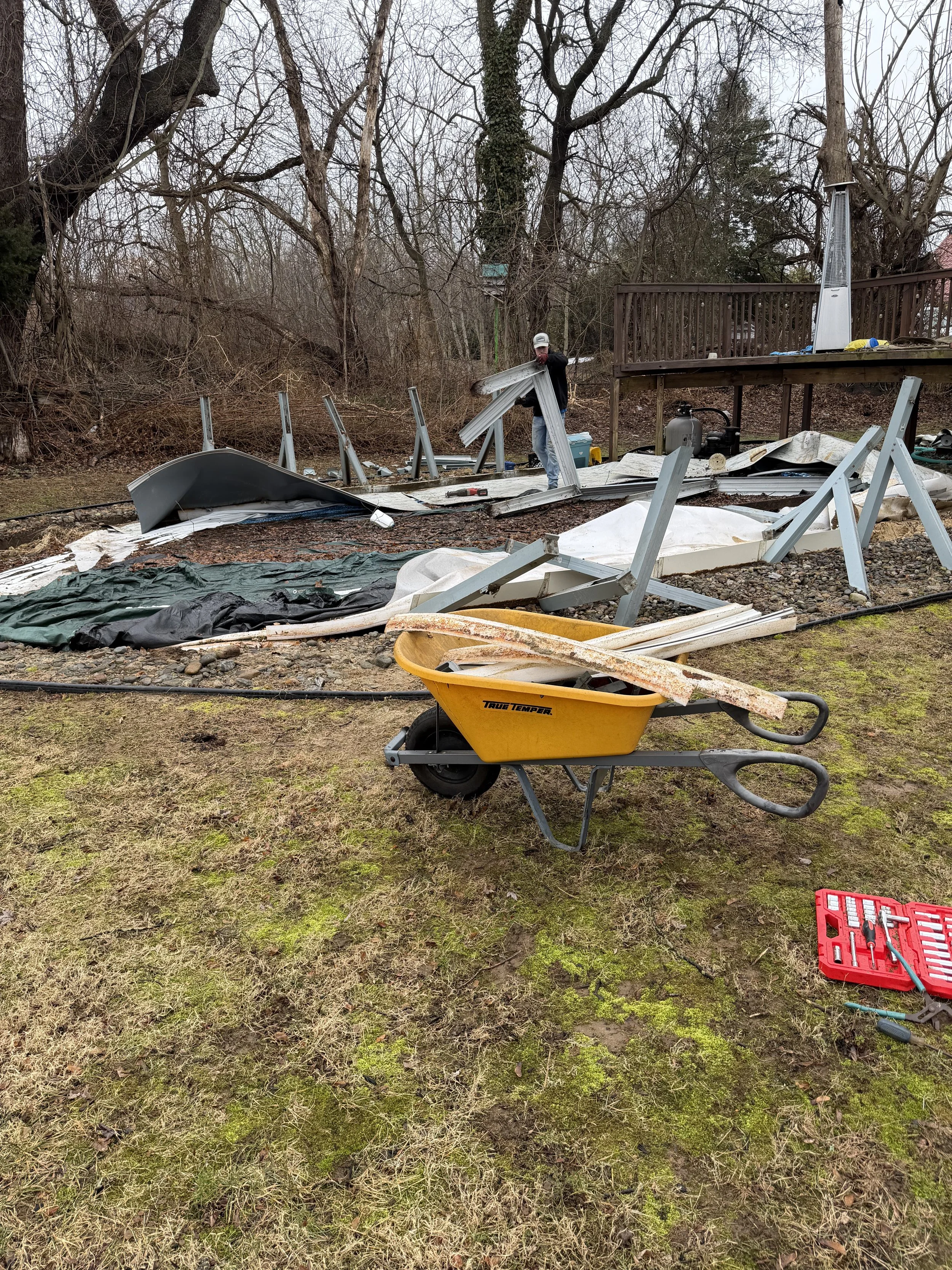 A person working outdoors on a broken structure, with metal poles and debris scattered around, a yellow wheelbarrow with rusted parts in the foreground, and tools on the ground nearby.