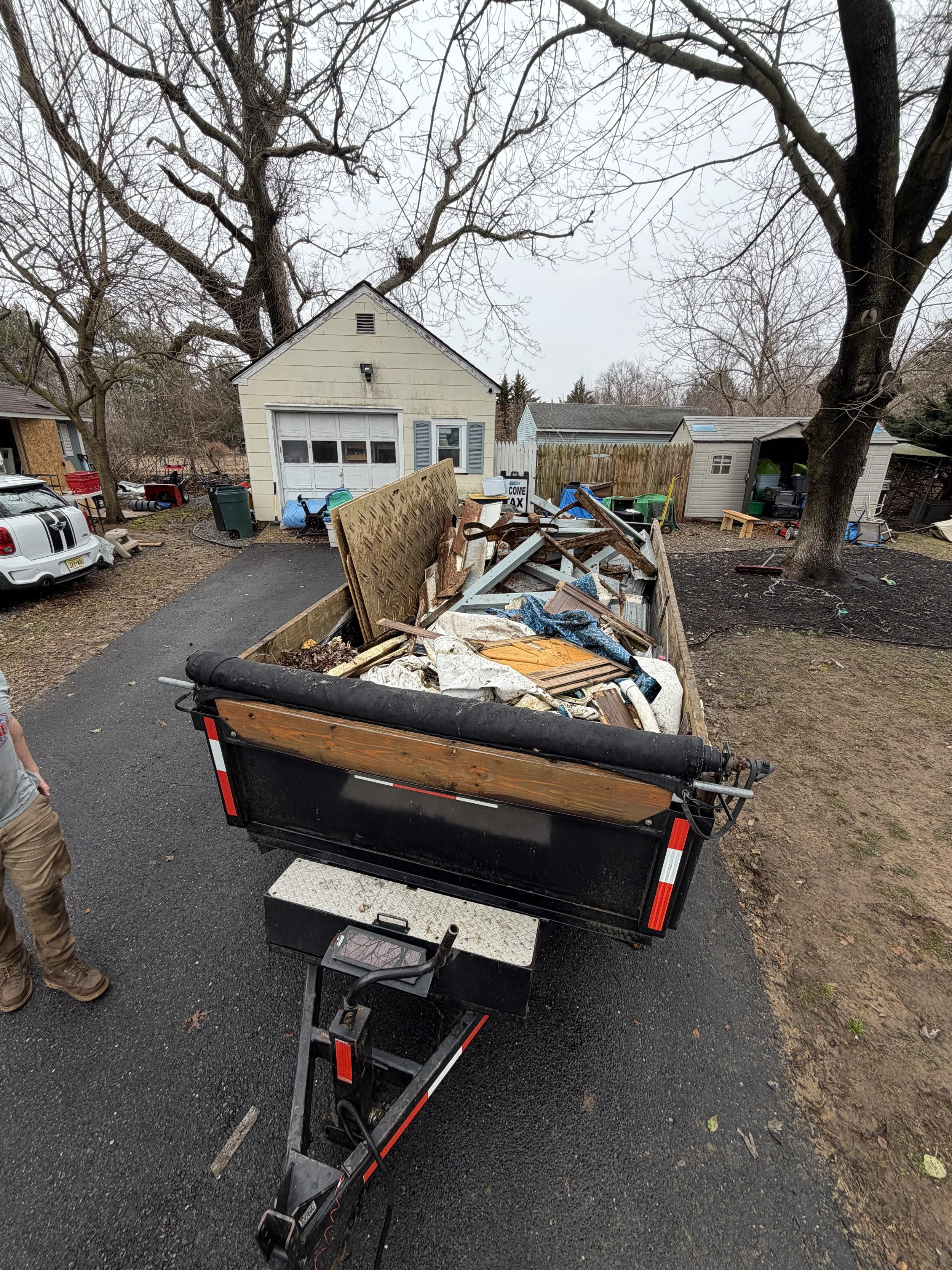 A trailer filled with discarded debris, including wood, metal, and trash, parked on a driveway in a residential backyard with leafless trees and various sheds.