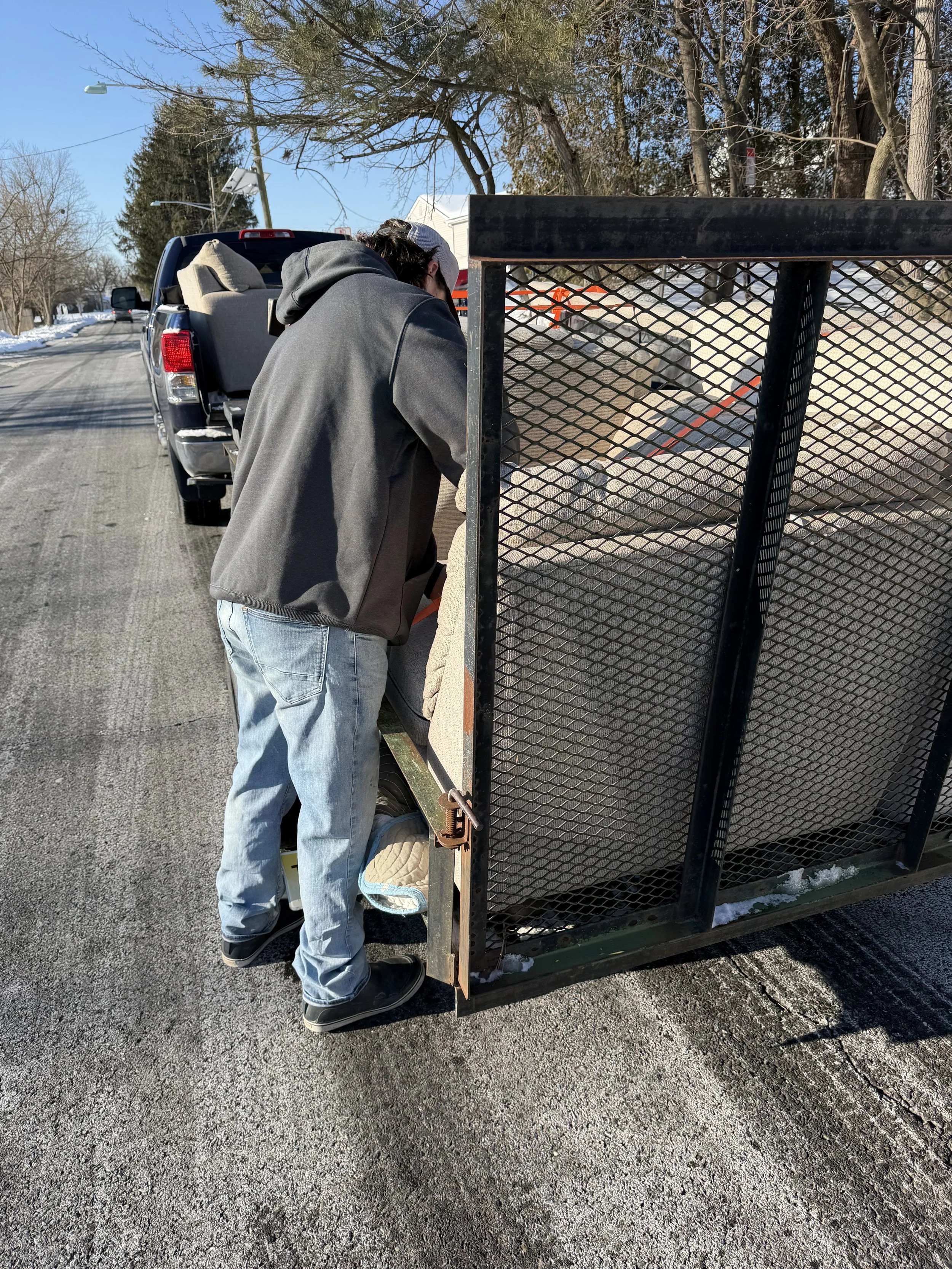 Two people loading or unloading items into a trailer behind a pickup truck on a residential street on a cold winter day.