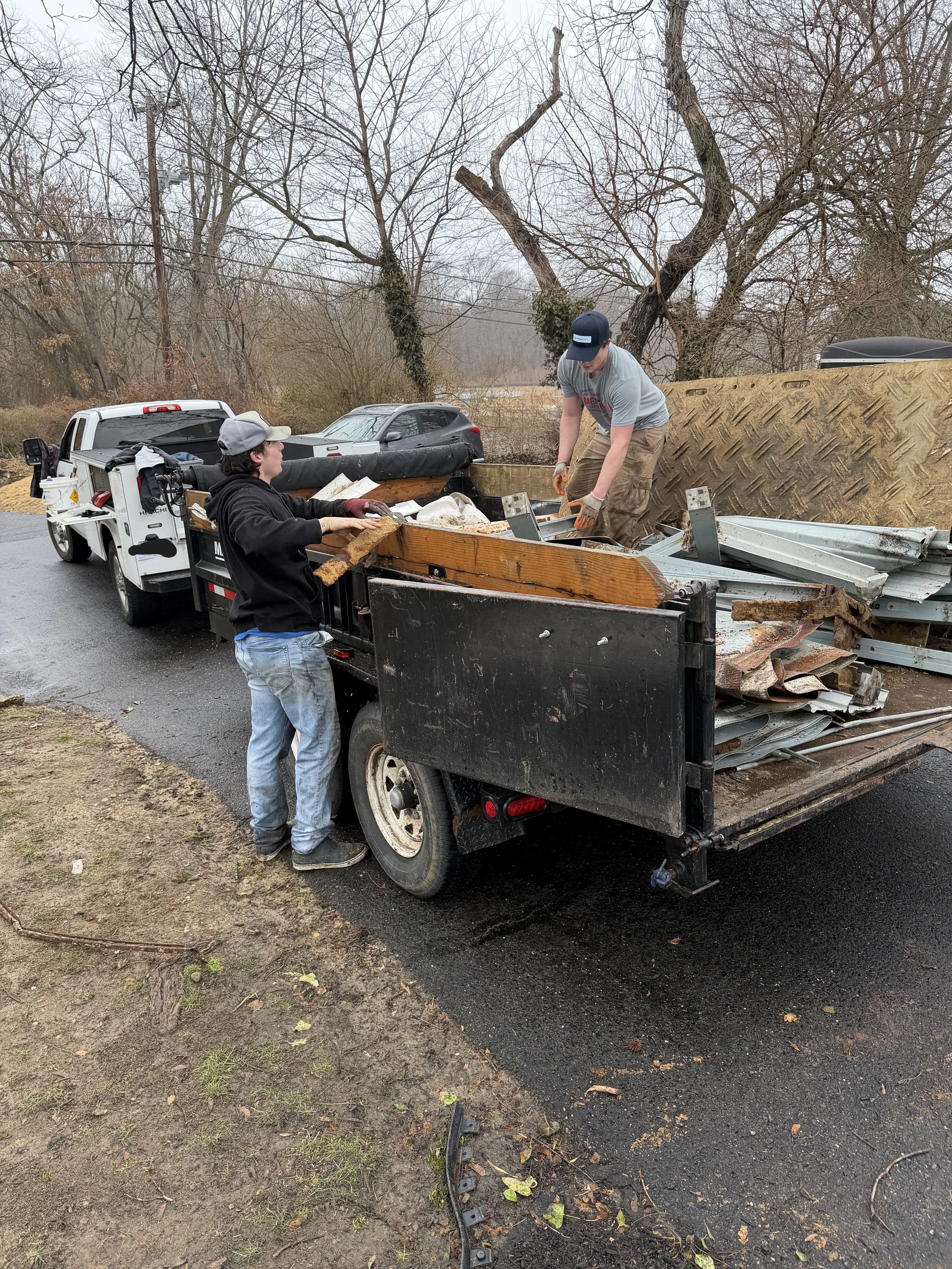Two people loading scrap metal and wood onto a flatbed truck parked on a wet road. One person is standing on the truck bed, and the other is beside the truck, passing materials.