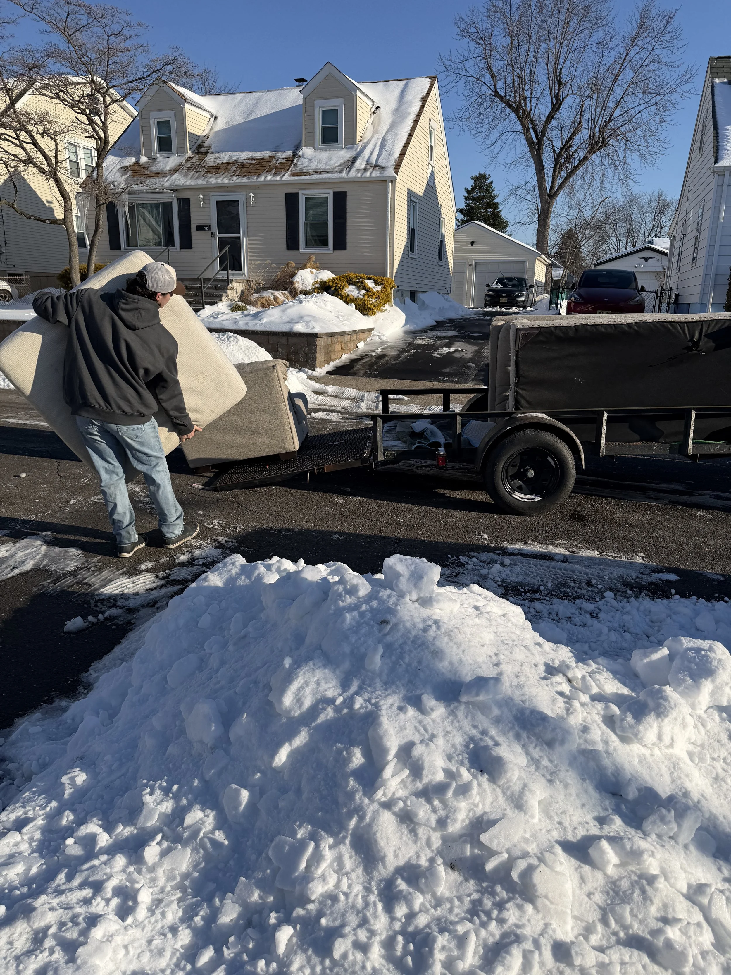 A man is loading a large beige sofa onto a trailer in a snowy residential neighborhood. The house in the background has a snow-covered roof and a small porch. There are snow piles on the ground and a clear blue sky.