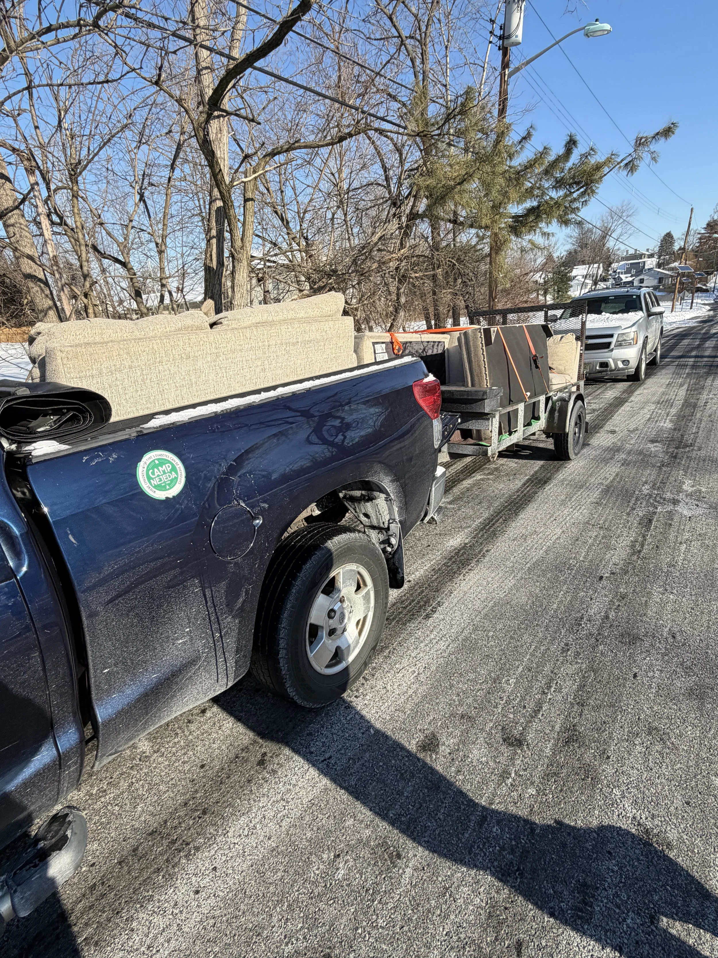 A dark blue pickup truck with furniture and boxes loaded in the truck bed and on a trailer attached to it, parked on a snowy street in a residential neighborhood with leafless trees and other vehicles in the background.
