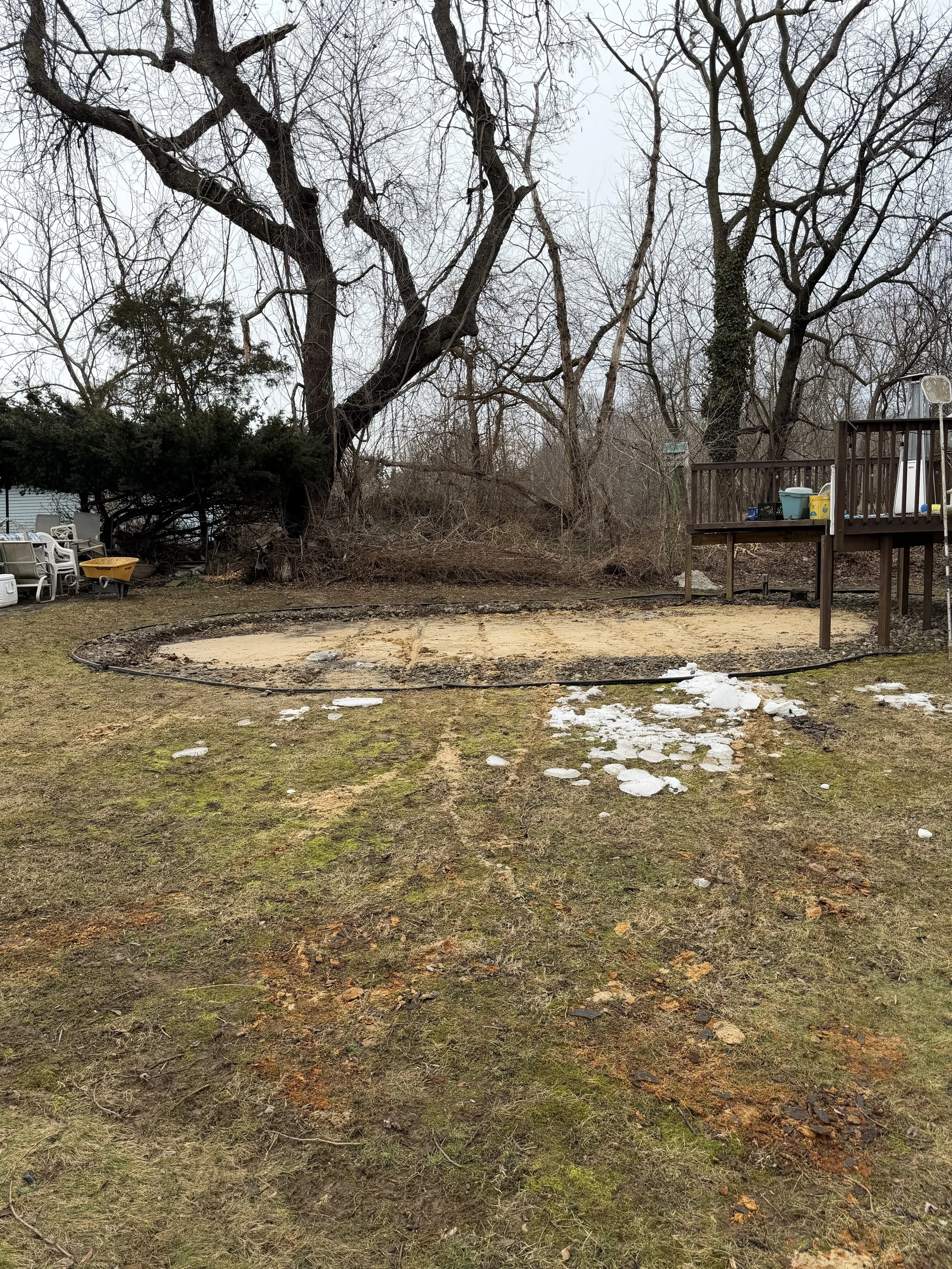 Backyard with a large, leafless tree, a wooden deck with stored items, and patches of snow and muddy ground.