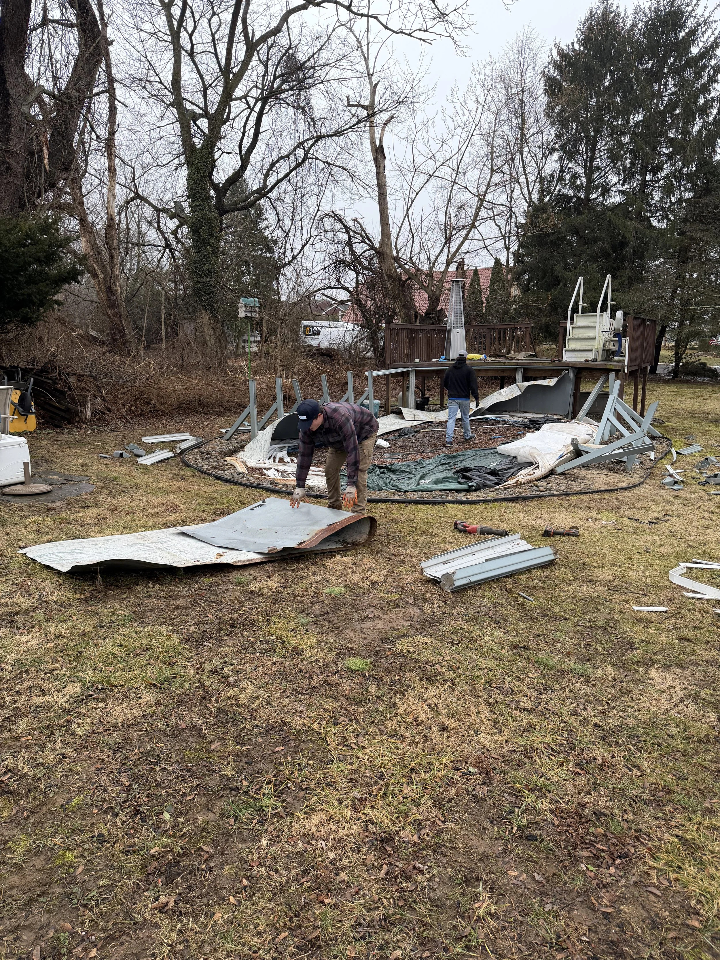 Two men dismantling a damaged above-ground swimming pool, with broken panels and debris scattered across the yard on a cloudy day.