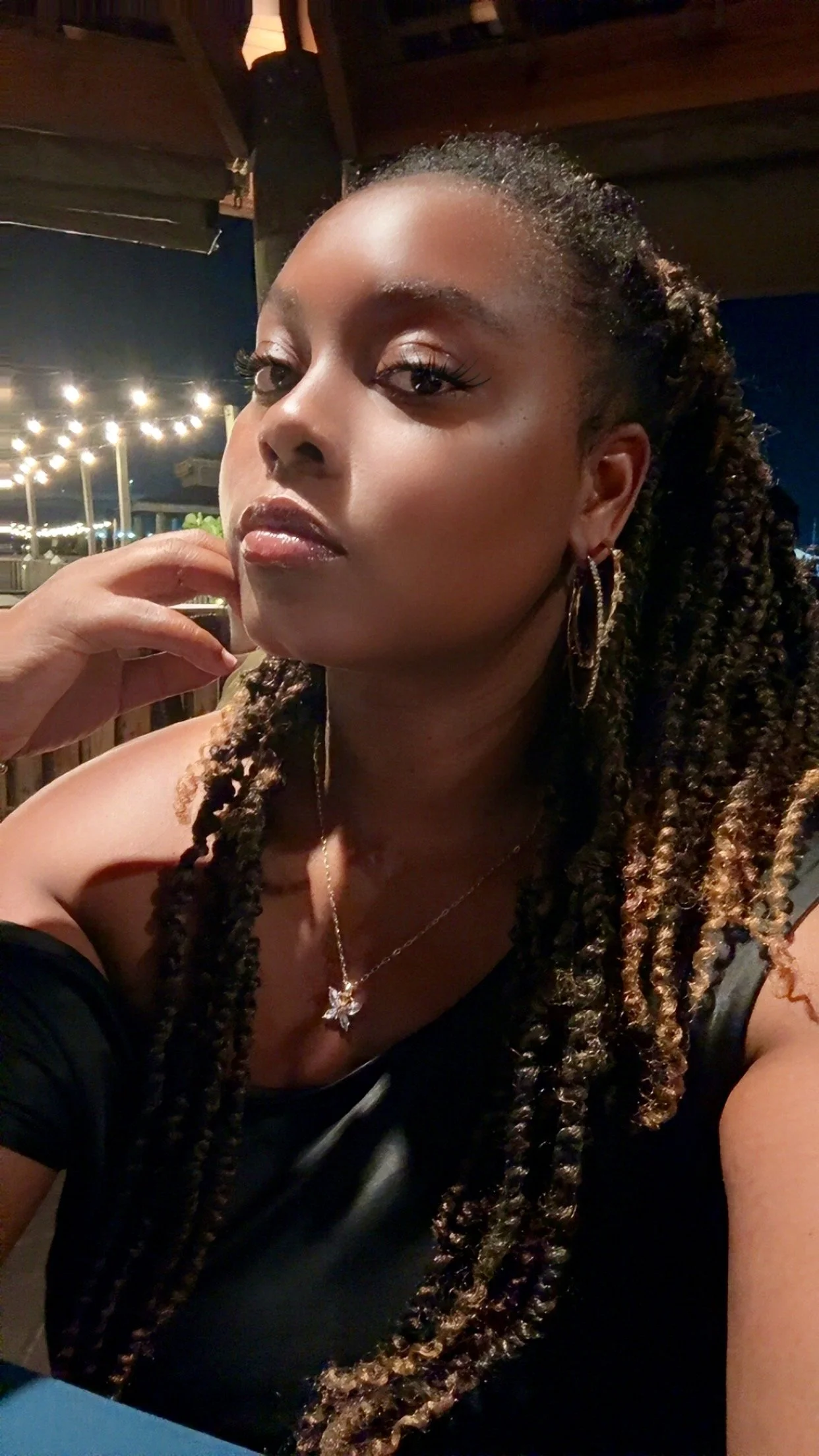 A woman with long braided hair, gold hoop earrings, a butterfly necklace, sitting outdoors at night under string lights.