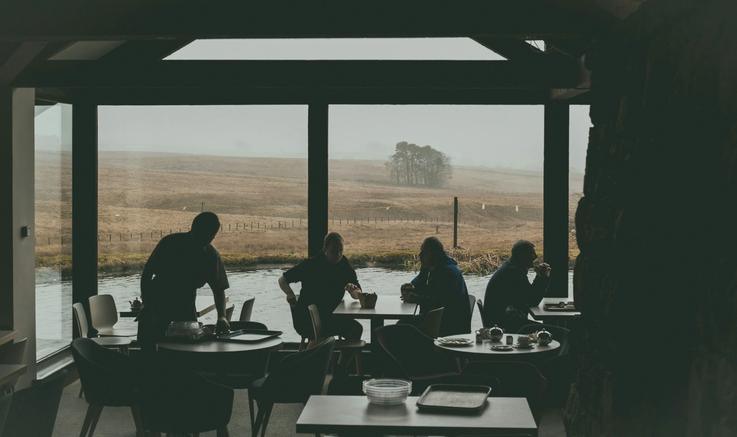 People sitting and standing around tables inside a restaurant with large windows overlooking a foggy, rural landscape with trees and hills.
