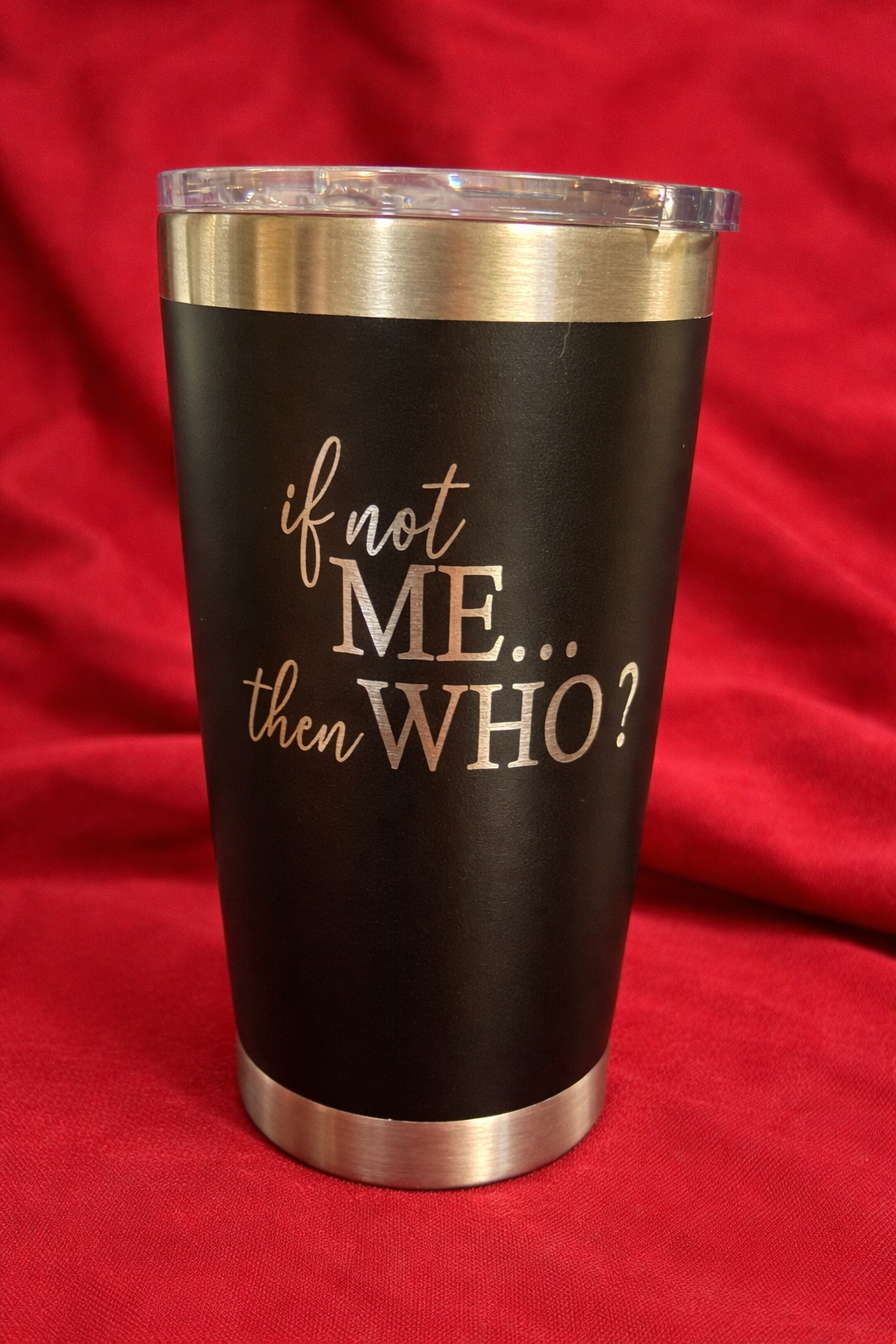 A black travel mug with silver trim and clear lid, featuring the text "if not ME... then WHO?" in white and gray lettering, set against a red fabric background.